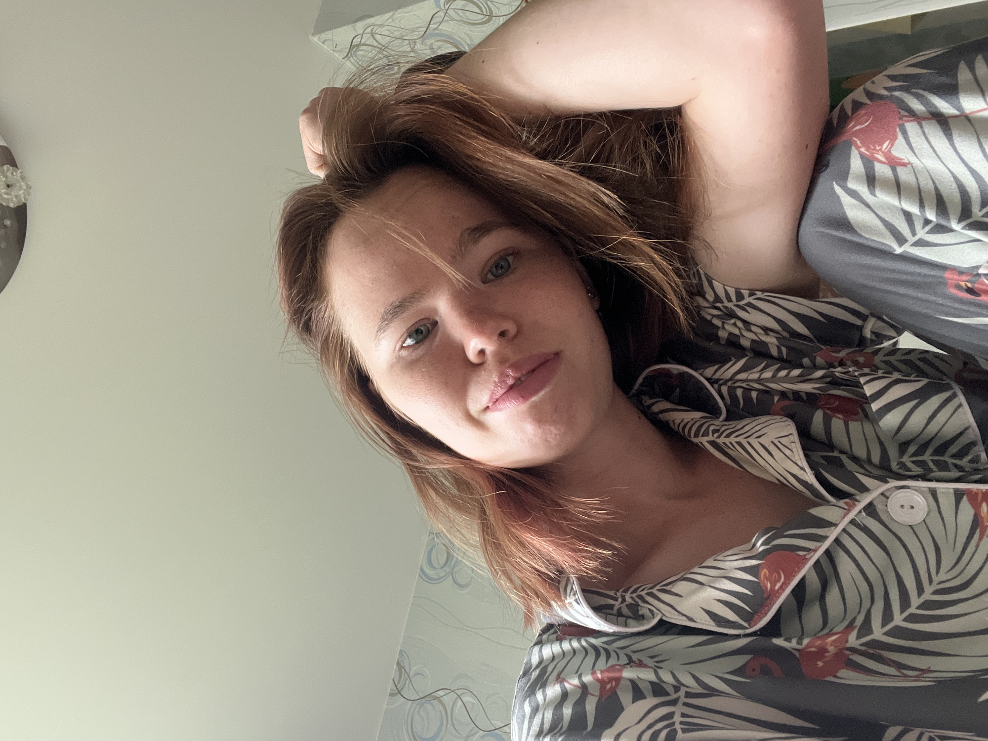 A low-angle, close-up, indoor, selfie shows a young woman with reddish-brown hair and blue eyes. She is wearing a pajama set with a tropical leaf and flamingo pattern. The woman is leaning her head against her right arm, which is bent at the elbow, and her left hand is resting on her head. Her eyes are looking towards the camera, and her lips are slightly parted in a pout. The background shows a light-colored ceiling with a decorative light fixture and a wall with patterned wallpaper. The overall impression is casual and relaxed, possibly taken in a bedroom in Chaikovsky, Russia.