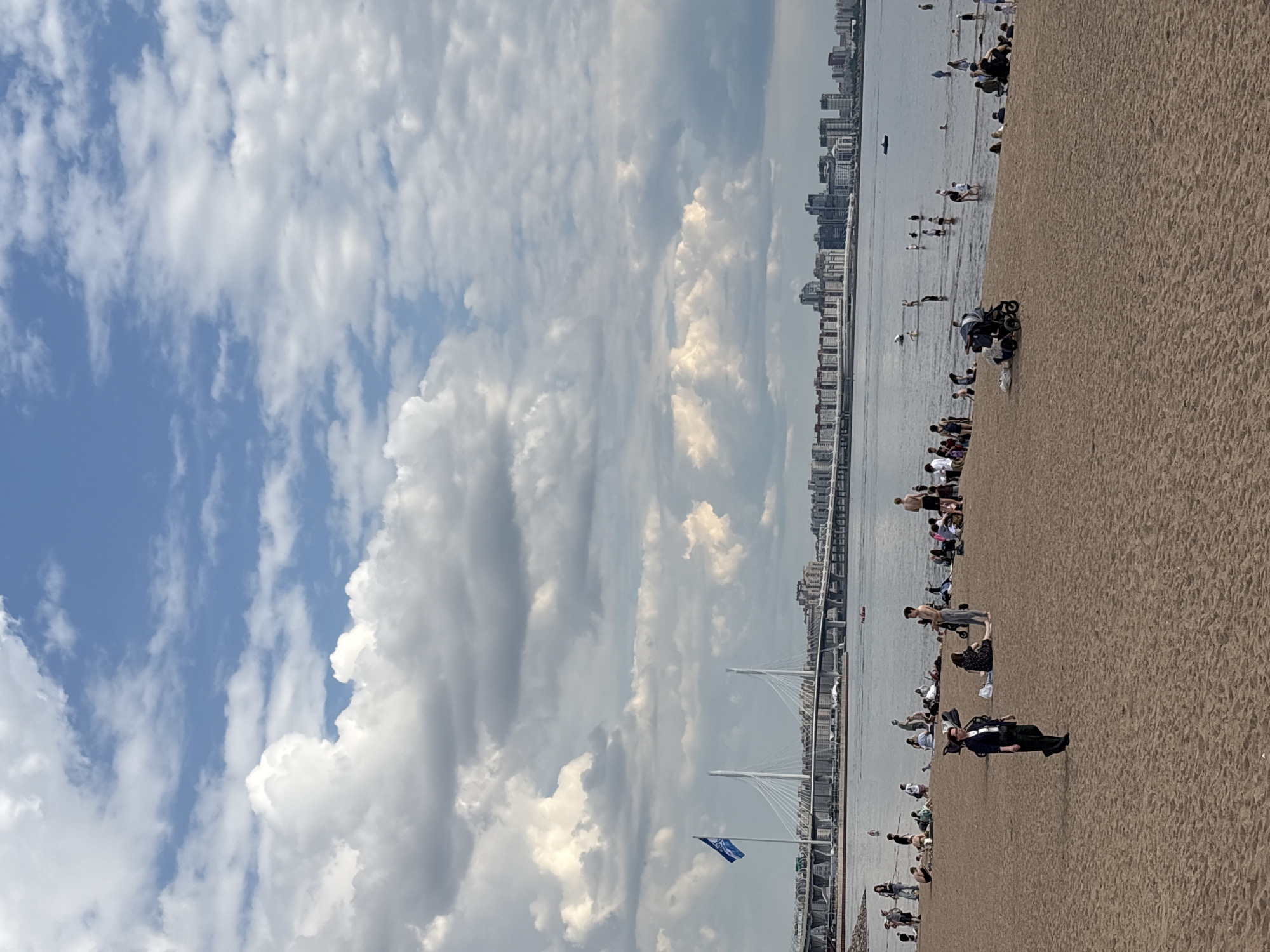 The image captures a bustling beach scene under a partly cloudy sky, likely in Saint Petersburg, Russia, given the context. The horizon is dominated by a wide body of water, possibly a bay or gulf, bordered by a distant city skyline featuring numerous buildings. Spanning across the water, a prominent modern cable-stayed bridge with white pylons adds an architectural element to the landscape.
In the foreground, a sandy beach is filled with numerous people. Many individuals are scattered across the sand, some sitting or standing, while others wade in the shallow waters along the shoreline. Activities include casual strolling, relaxing on the sand, and enjoying the water. One person is pushing a baby stroller on the sand, and another person dressed in black stands prominently.
The sky above is a mix of bright blue patches and large, fluffy white and grey clouds, suggesting a partly sunny day. The lighting is bright but somewhat diffused, indicating it is daytime, possibly late morning or afternoon. A blue flag with a white emblem or logo is visible on a pole near the bridge, fluttering in the breeze. The overall impression is one of leisure and outdoor recreation on a sizable urban beach.