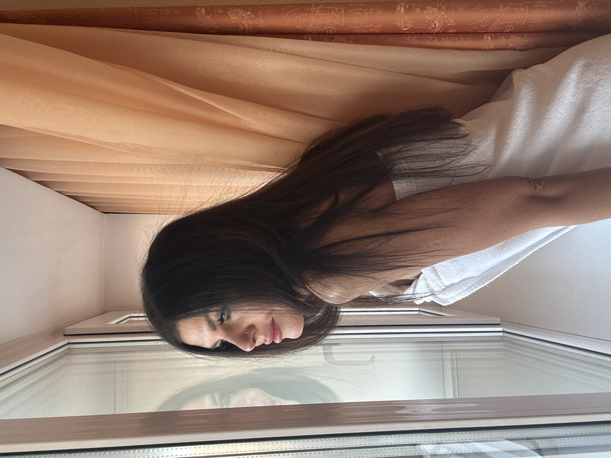The image features a young woman with long, dark hair, captured in an indoor setting. She is positioned looking downwards and to her left, with a pensive or contemplative expression, her lips slightly parted. A small, metallic piercing is visible on her right nostril. Her dark hair cascades over her shoulders and upper back.
She appears to be wrapped in a white, soft garment, possibly a towel, which is visible around her upper body and right arm. The woman is standing next to a modern white-framed window, and her reflection, including her face and hair, is clearly visible on the glass pane below her.
In the background, light peach or orange sheer curtains are drawn, creating gentle folds. Behind these, a darker, patterned fabric, likely another curtain, adds texture to the upper part of the scene. The walls in the room are plain and light-colored. The presence of natural light suggests it is daytime. The overall scene conveys a quiet, personal moment, possibly post-shower or during a moment of introspection. No text is visible in the image.
