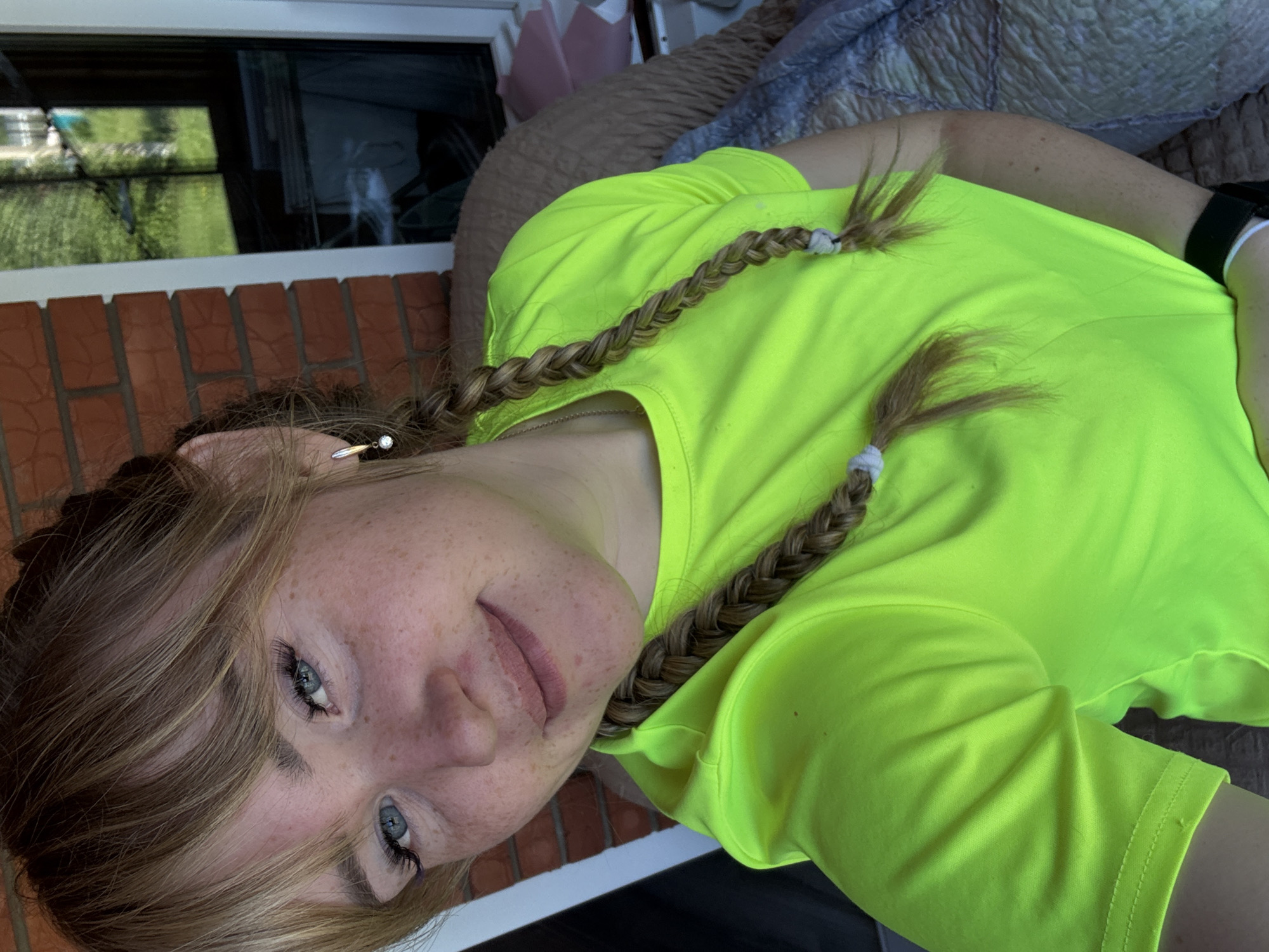 A young adult female is depicted in a close-up, chest-up selfie taken in Ляльшур, Russia. She has light skin, prominent freckles across her nose and cheeks, and light blue eyes with dark eyelashes. Her light brown hair is styled with bangs and two braids, which fall forward over a bright neon green short-sleeved t-shirt. She is wearing a small gold-toned earring with a clear stone in her right ear and a dark wristband with a white accent on her left wrist. The immediate background behind her head is a reddish-brown brick wall. To her right, a white window frame reveals outdoor greenery and a distant structure. To her left, a textured, brown upholstered surface and a light blue patterned blanket are visible. She is looking directly at the camera with a slight smile.