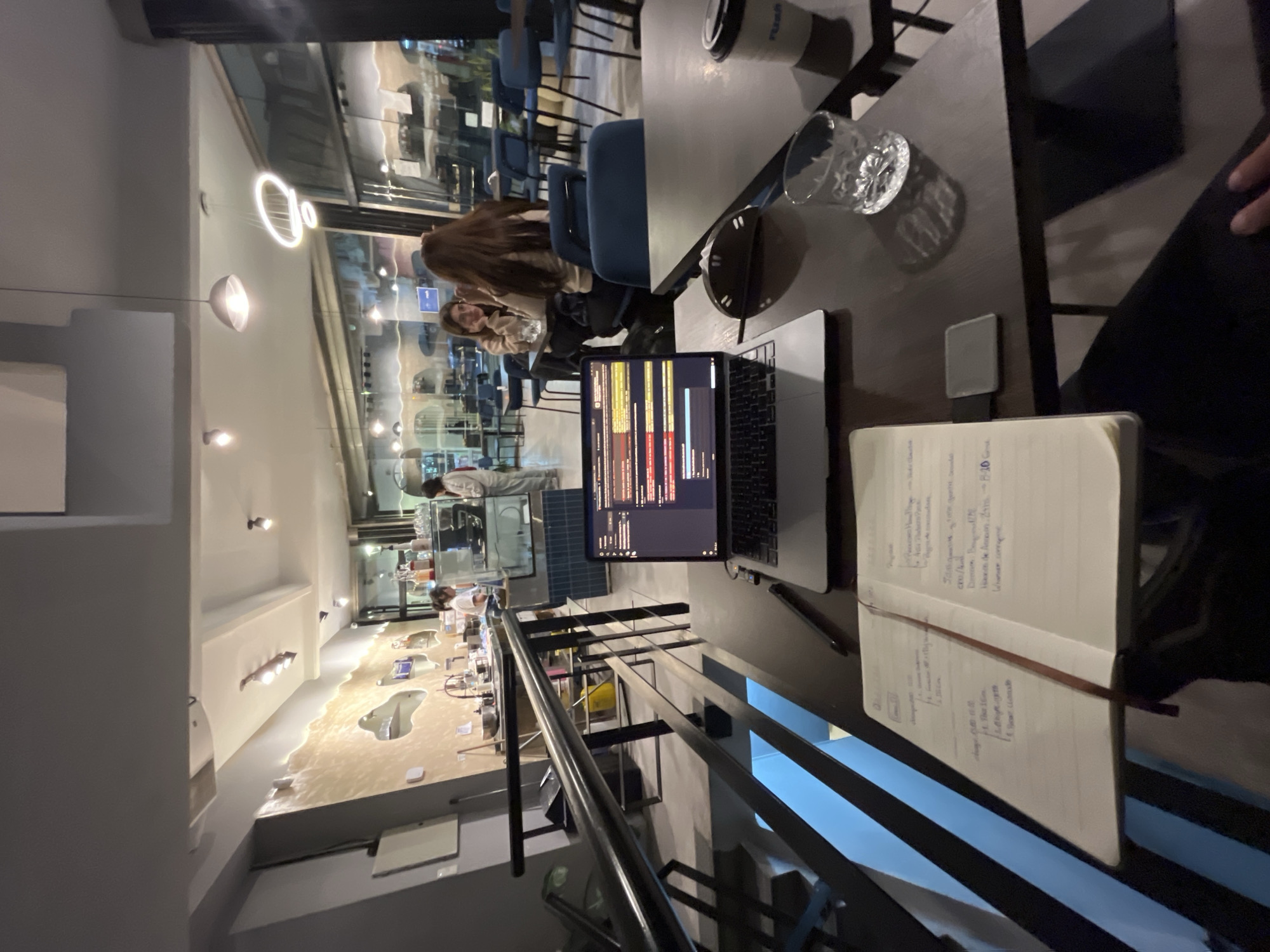 This image captures an indoor scene, likely a cafe or co-working space, at night or in the evening. The perspective is from a high-angle, looking down slightly onto a dark-colored table in the foreground and into the establishment.
In the immediate foreground, a silver laptop is open, displaying lines of code with various syntax highlighting colors on its screen. To the right of the laptop, an open notebook with handwritten text is visible, held down by a dark bookmark ribbon. A clear glass containing water, a small silver rectangular object (possibly a card reader or portable charger), and a partially visible white coffee cup with a blue rim (brand name "Purav" or similar) are also on the table. A hand, presumably the photographer's, rests on the table's edge near the notebook.
In the mid-ground, two women are seated at a table. One has long, dark brown hair and is looking towards the other, who has shorter hair and is looking slightly towards the camera with a subtle smile. They are surrounded by dark blue chairs and other tables.
The background reveals a spacious interior with modern lighting fixtures, including a large circular light and bowl-shaped pendants. Beyond the seating area, there is a counter where staff members are visible, suggesting a service area. A distinctive patterned wall or floor design, resembling a sandy landscape or a topographical map, adds visual interest to the space. Reflections in large windows or glass partitions indicate it is dark outside. Stairs with black metal railings lead downwards in the lower-left section of the image.
The activity suggests a combination of work/study and casual social interaction. The laptop user is engaged in coding or similar work, while the women in the mid-ground appear to be conversing or relaxing.
Visible text in the open notebook includes headings like "Description," "Requirements," "Goals," and "Next Steps." Crucially, the location "Buenos Aires, Argentina" is clearly written on the right page, along with "P-20," "Garruk," "Amazon," and phrases like "dir. of all departments" and "Urbanas campagnes."