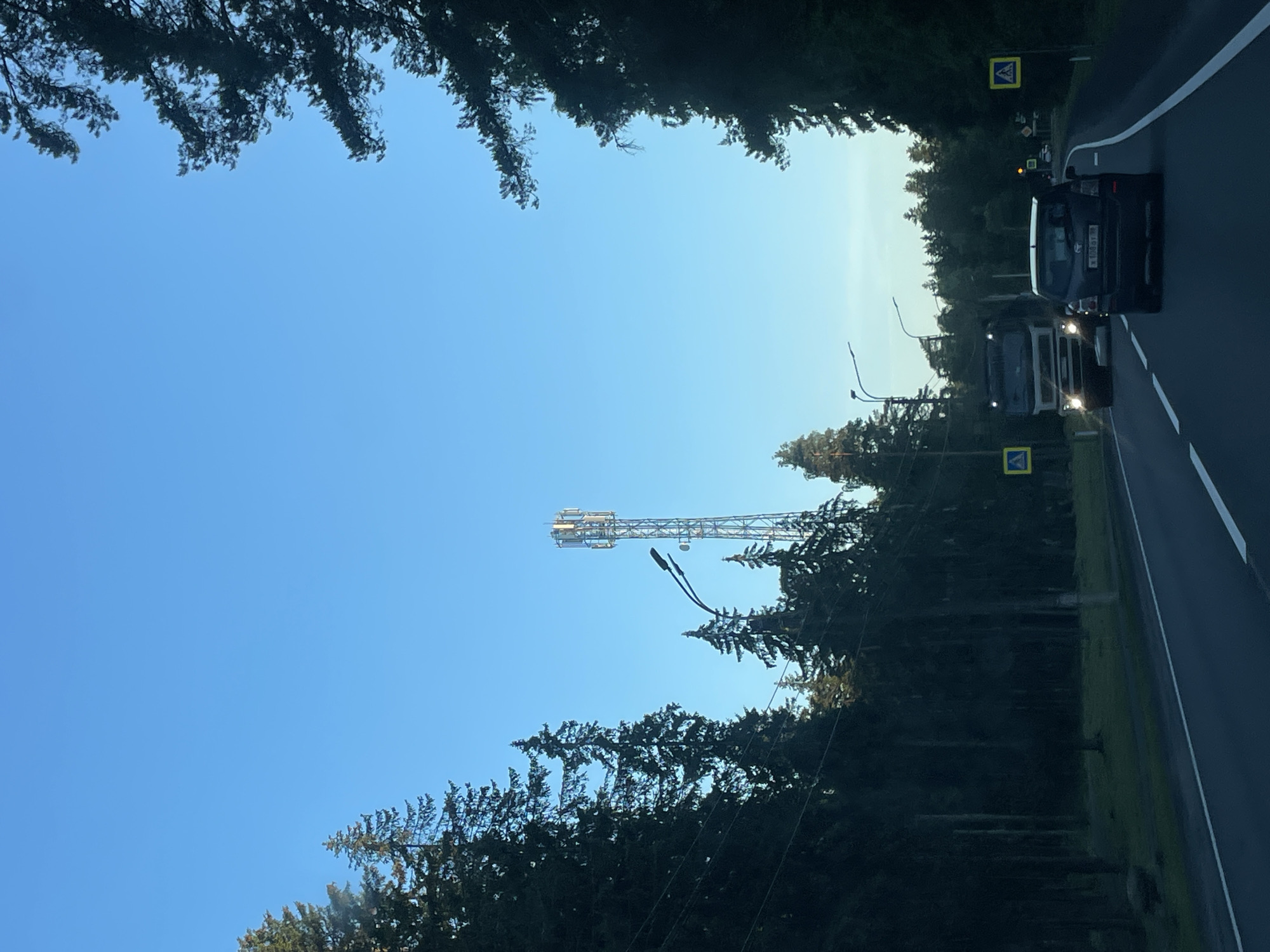 The image captures a road scene in Smolyachkovo, Russia, under a clear blue sky. A tall telecommunications tower is visible in the distance, situated amongst a dense forest of evergreen trees. The road itself is paved with white lane markings and is bordered by the dark silhouettes of trees on both sides. A white truck with its headlights on is driving on the left side of the road, while a dark-colored car is following behind it. Yellow pedestrian crossing signs with a blue pedestrian icon are posted on the right side of the road. The overall environment suggests a rural or semi-rural setting with a road passing through a forested area.