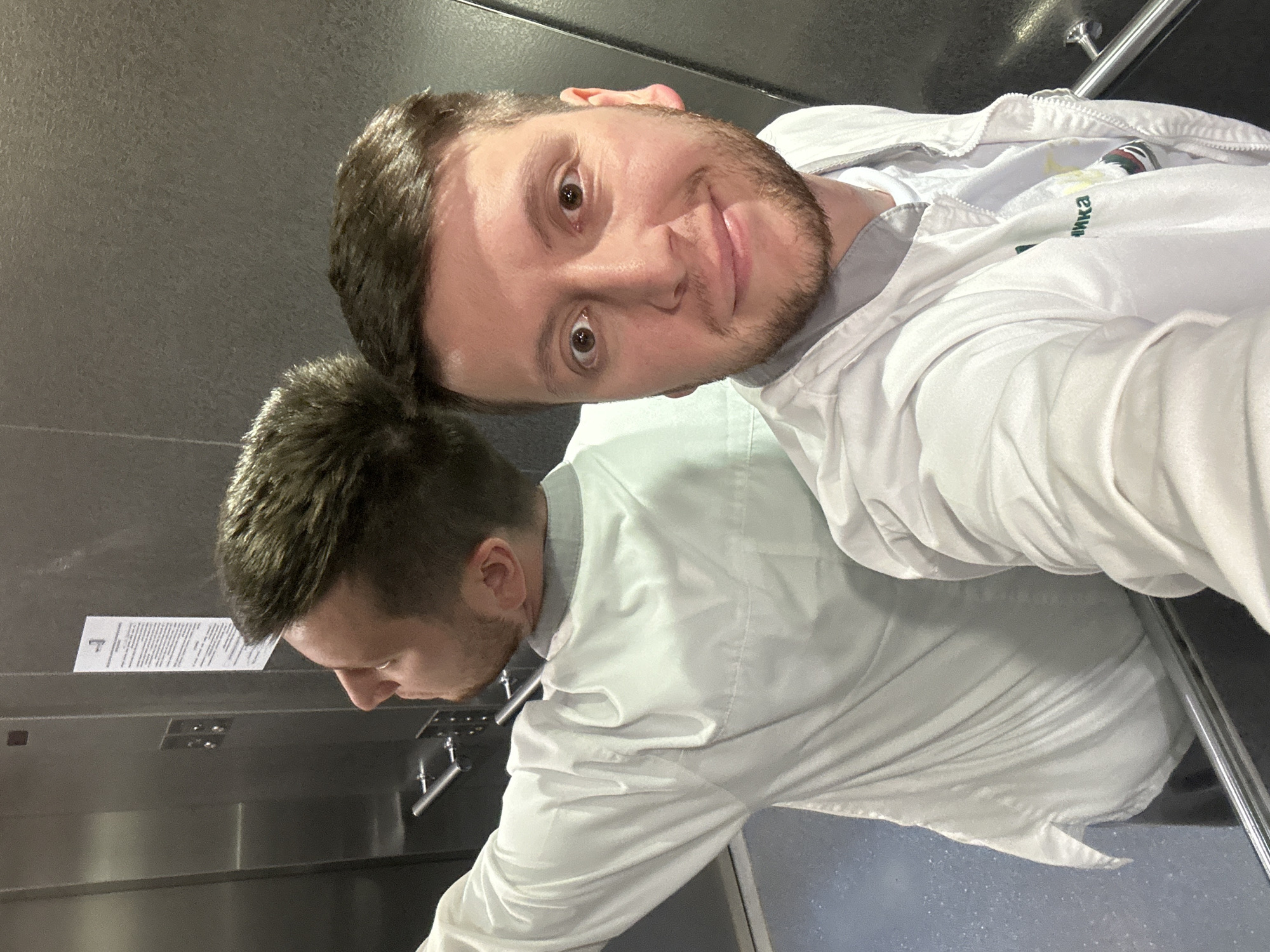 A male individual is captured in a selfie taken inside an elevator. He is a young adult with light brown hair and a beard, wearing a white zip-up jacket, possibly a medical scrub or lab coat, over a white undershirt. Part of a green Russian word, "ика," is visible on the jacket's front left side, suggesting "клиника" (klinika), meaning "clinic" or "hospital." His eyes are wide, and he exhibits a slight smile. The man's reflection is prominently visible behind him in the elevator's polished metallic wall, showing him from the back and left profile, seemingly looking towards the elevator's control panel. The elevator interior features metallic walls, a control panel with multiple buttons (square and round), and a silver-colored handrail on the right. A white paper notice with black text is affixed to the wall, visible in the reflection. The floor appears to be a light blue or gray speckled surface. The scene depicts a medical professional in an elevator, likely within a hospital or clinic setting in Moscow, Russia.