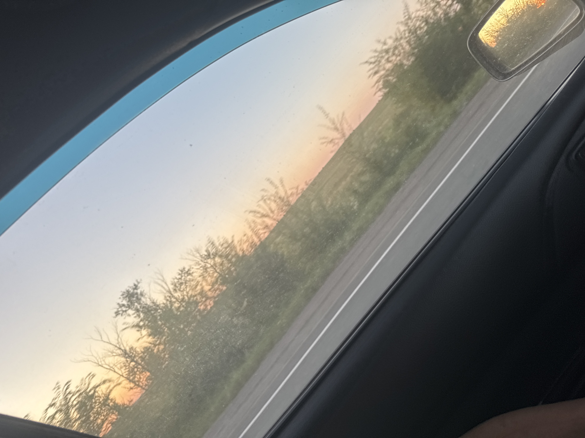 The image shows a view from inside a vehicle looking out a window towards a sunset. The sky is a gradient of light blue at the top, fading into pale orange and pink near the horizon. Silhouetted trees and brush line the side of a road, which is visible in the foreground. The road has a white line marking its center. A side mirror of the vehicle is visible on the right side of the frame, reflecting the sunset and trees. The interior of the vehicle, including a portion of a door panel and an arm, is visible in the lower part of the image. The image appears to be taken in Ostrolenskii, Russia, during a sunset.