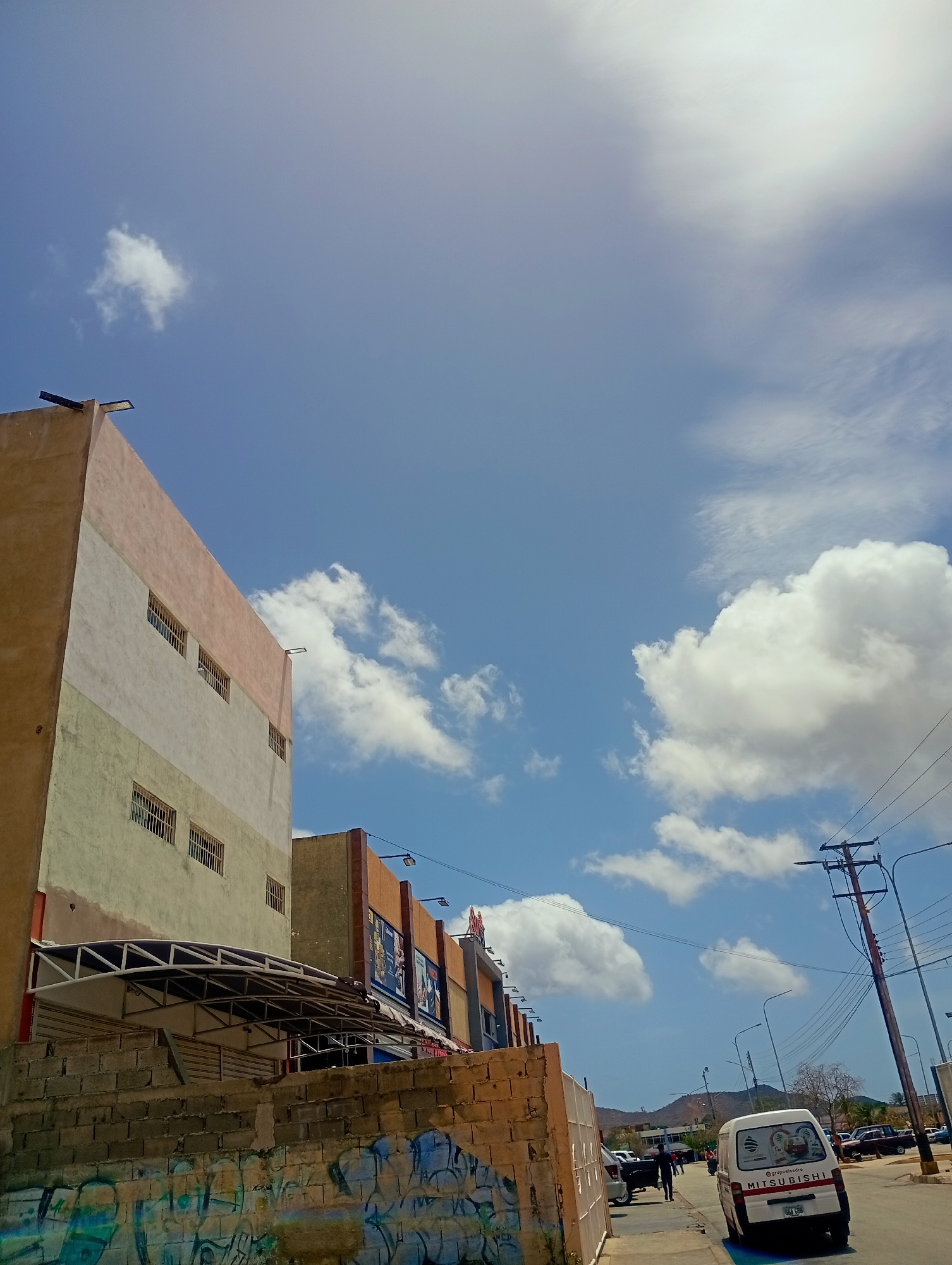 A wide shot captures a street scene in Juan Griego, Venezuela, under a bright blue sky dotted with white clouds. In the foreground, a graffiti-covered brick wall runs horizontally across the bottom of the frame. Behind the wall, a building with a multi-colored facade, featuring horizontal bands of beige, pale green, and pink, rises on the left. This building has several rectangular windows with metal grates. A white awning with a dark metal frame extends from the front of the building over a walkway. To the right of this building, another building with an orange and brown facade is visible, displaying posters and signage. A white Mitsubishi van is parked on the right side of the street, facing away from the camera. Several other vehicles, including cars and a pickup truck, are parked or driving further down the street. A utility pole with multiple wires stands on the right, alongside streetlights. In the background, a line of hills or mountains is visible against the horizon. The overall scene depicts a commercial or industrial area on a sunny day.