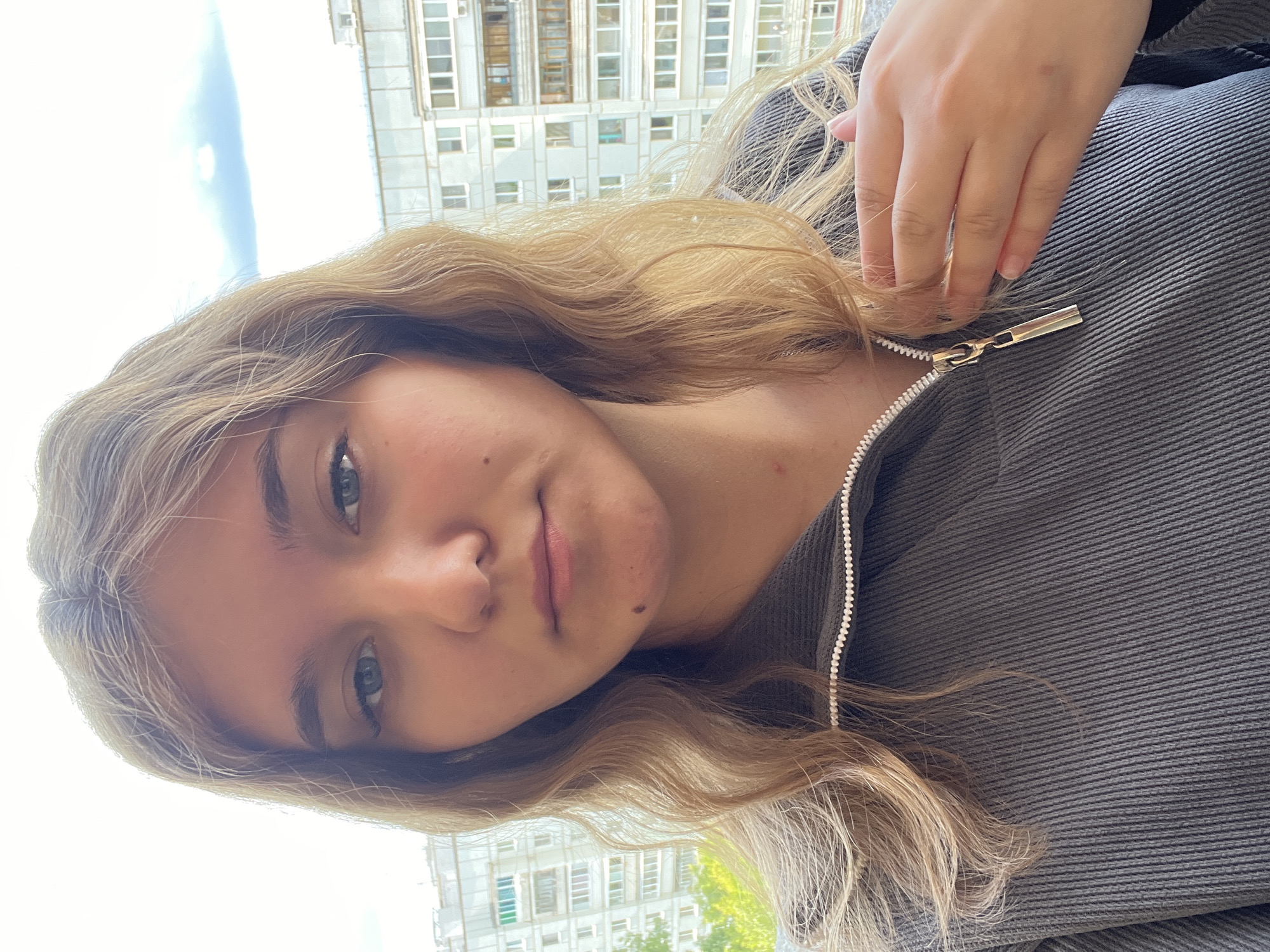 A young Caucasian woman with shoulder-length blonde hair and blue eyes is pictured in a close-up, waist-up shot. She is wearing a dark grey zippered jacket. Her left hand is placed on her chest, near the zipper. The background shows a residential building with multiple floors and windows, characteristic of the city of Chaikovsky, Russia. The sky is visible above the building, with scattered clouds. The lighting suggests it is daytime, possibly late afternoon due to the soft quality of the light. The woman appears to be looking directly at the camera. There are no other people or animals visible in the image. The environment appears to be outdoors, on a balcony or near a window with a view of the urban landscape.