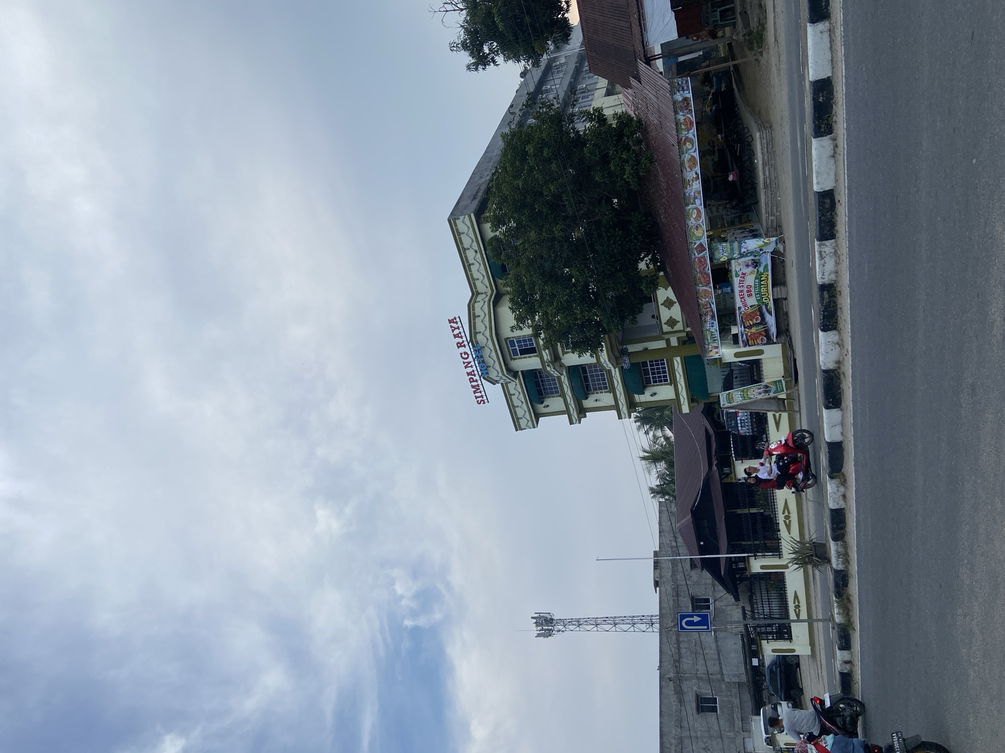 This is a street-level, medium-shot photograph taken in Tanjung Gading, Indonesia, during the day under an overcast sky. In the background, a multi-story building with a green and cream facade is visible, bearing the red signage "SIMPANG RAYA HOTEL" at the top. Adjacent to the hotel, a covered outdoor seating area with tables and chairs is set up in front of what appears to be a food stall. Various food advertisements are displayed along the front of this area. A cellular tower stands tall in the mid-ground to the left of the hotel. In the foreground, a two-lane asphalt road is visible, separated by a raised median with black and white markings. Several motorcycles are on the road. A red scooter carrying two people, a male driver and a female passenger, is in motion on the right side of the road. Another motorcycle with a single rider is in motion on the left side of the road, closer to the foreground. Behind the median, a dark-colored building with a sloped roof is situated, and a black metal fence encloses a property. Further to the left, a concrete building with rectangular windows is present. A blue U-turn traffic sign is mounted on a pole near the concrete building. The overall environment suggests a commercial or roadside area with ongoing traffic and activity.