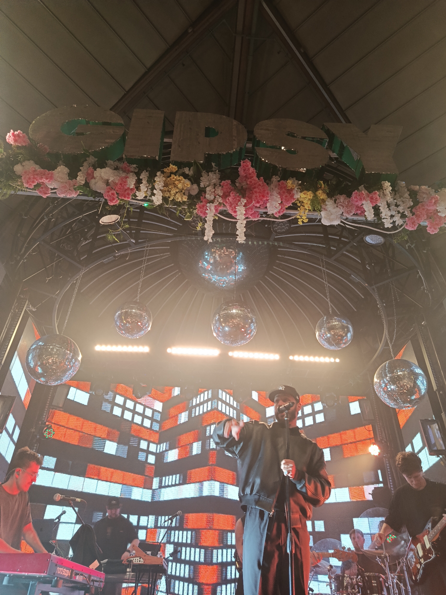 The image depicts a live musical performance in an indoor venue in the city of Unknown, Unknown. Above the stage, a large sign spelling "GIPSY" is adorned with pink and white flowers. Several disco balls hang from the ceiling. On stage, a male vocalist wearing a black jacket, black trousers, and a baseball cap is front and center, holding a microphone and gesturing with his right hand. Behind him, a large LED screen displays an orange and white block pattern resembling a cityscape. Other band members are visible playing instruments: a keyboard player is on the left, a guitar player on the right, and a drummer is positioned further back in the center. Additional musicians are partially visible in the background. The lighting creates a bright and energetic atmosphere.