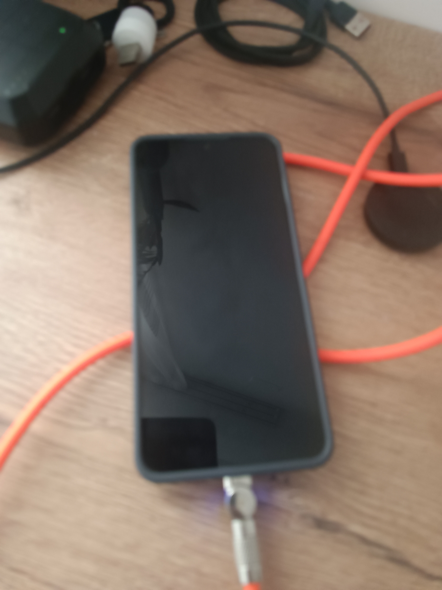An out-of-focus, high-angle shot shows a black smartphone connected to an orange charging cable. The phone is lying horizontally on a wooden surface. To the left of the phone, a black electronic device with a green light is partially visible. In the background, a black cable with a USB connector is also present. The scene is likely an indoor setting, possibly a desk or table. The city of Silifke, Turkey, is not depicted or relevant to the image content.