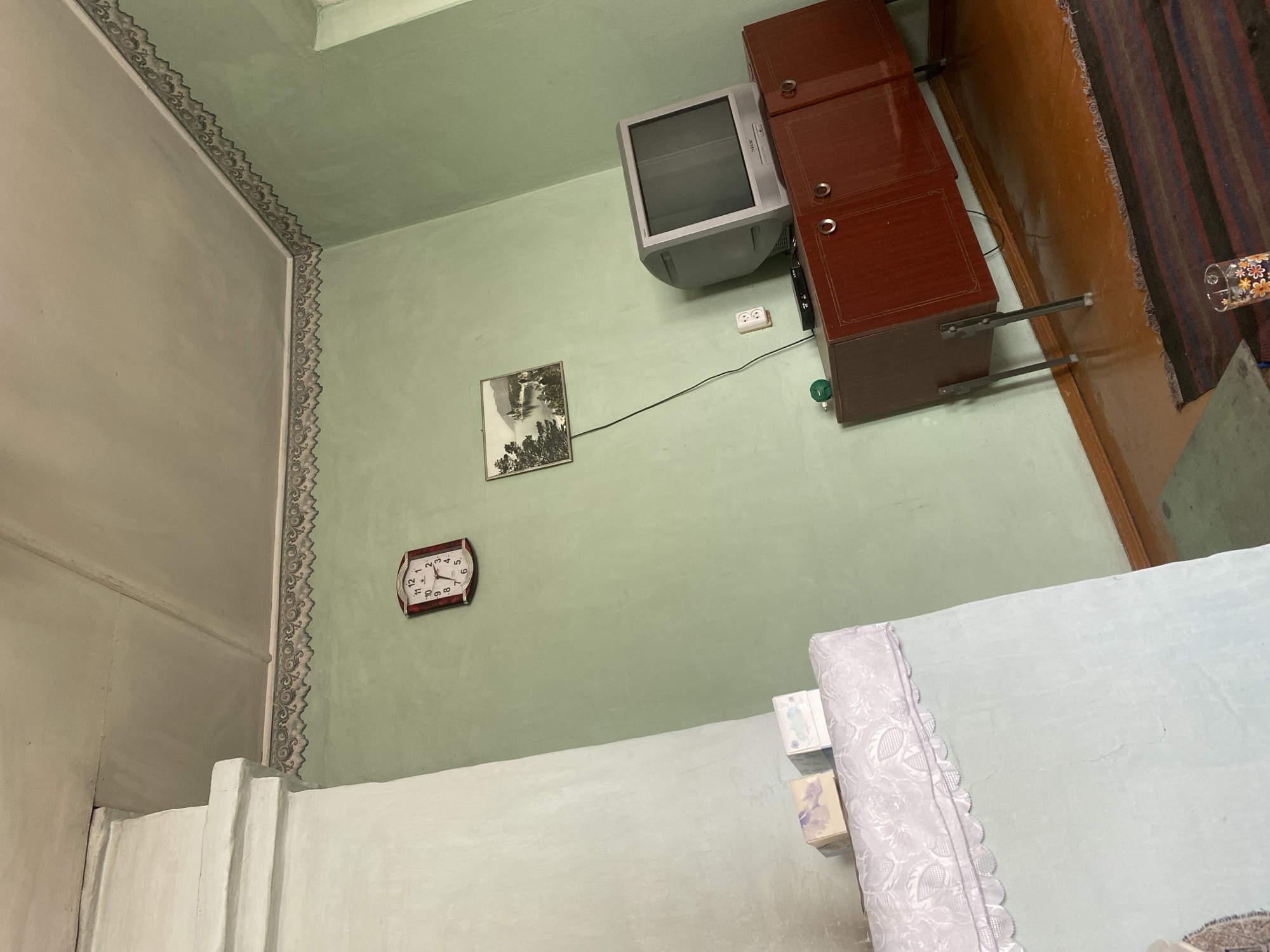 The image shows an interior view of a room, likely a living room, in Usolye-Sibirskoye, Russia. The walls are painted a light green, and the ceiling appears to be textured and light-colored. A decorative border runs along the top of the walls where they meet the ceiling.
On the left side of the image, a large, light-colored structure, possibly a stove or a decorative fireplace, occupies a significant portion of the frame. On top of this structure, two small decorative boxes are visible.
To the right of the stove, a rectangular wall clock with a dark frame and a white face is mounted on the wall. The clock has Roman numerals and is showing the time as approximately 3:25.
Further to the right, a framed picture hangs on the wall, depicting a landscape with water and trees. Below the picture, a vintage television set is placed on top of a dark wooden cabinet. The television is a CRT model with a silver-grey casing. The cabinet has two doors with round, metallic handles. There is also a small green object on top of the cabinet, next to the television. An electrical outlet and a small black electronic device are visible on the wall and cabinet respectively.
The floor is made of polished wood, and a striped rug with dark red, brown, and black stripes is partially visible in the foreground. A glass with a floral pattern is placed on the rug.
The overall impression is of a lived-in, perhaps older, room. The lighting appears to be natural, coming from a window that is not fully visible on the right side of the image. There are no people visible in the image.