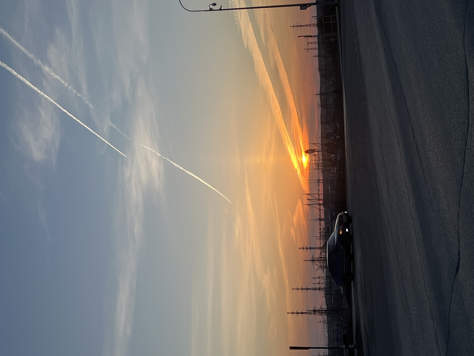 The image depicts a sunset in an industrial area, likely in Chaikovsky, Russia, given the presence of power lines and factory infrastructure. The sky transitions from blue to orange as the sun sets, creating a vivid display. Two prominent contrails streak across the blue part of the sky. A car is parked along a paved road, the only visible object at ground level. The overall environment seems quiet.