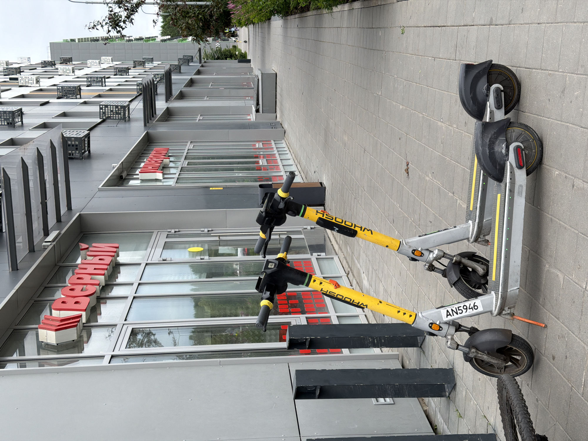 Two electric scooters, branded "WHOOSH," are parked on a paved sidewalk in front of a modern building. The building has large glass windows and a gray facade with red lettering that reads "Верный" (Verniy) in Russian. The scooters are yellow and silver, with black handlebars and wheels. One scooter has a license plate or identification number visible, "AN5946." To the left of the scooters, a bicycle wheel and frame are visible, suggesting another vehicle has been left there. The background shows a pathway leading away from the building, lined with greenery and more modern architecture. The scene appears to be in a well-lit, urban environment, possibly a commercial or residential area.