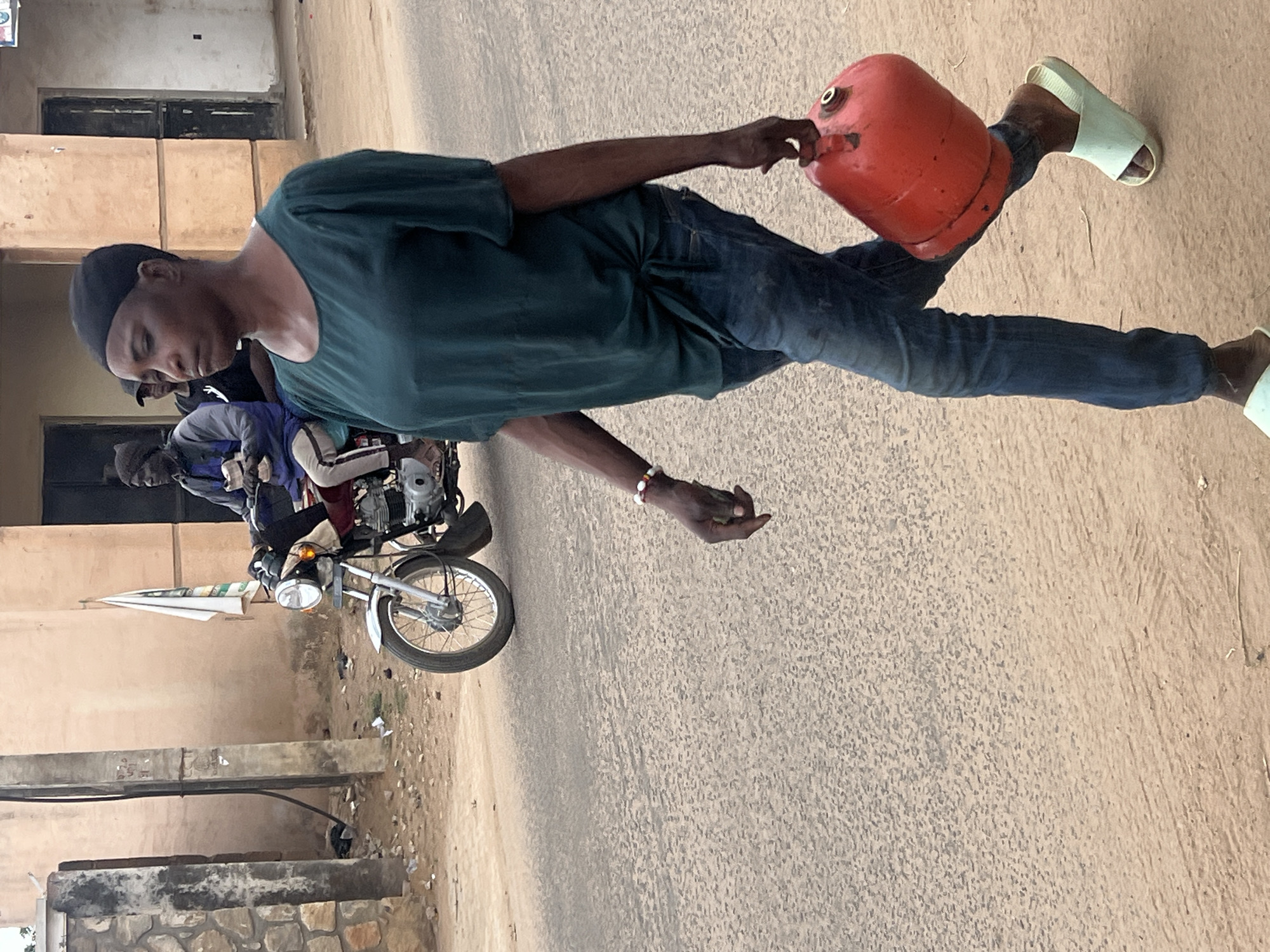 The image depicts an outdoor street scene, likely in Jos, Nigeria, focusing on a young man carrying a red gas cylinder. The main subject, a dark-skinned man, is seen from the waist up, wearing a black cap, a dark teal or deep green short-sleeved t-shirt, and blue jeans. He has light-colored sandals or slides on his feet and a multi-colored beaded bracelet on his right wrist. He holds the red, slightly worn gas cylinder by its top handle with his right hand as he walks, with his gaze directed downwards.
In the background, to the left of the main subject, two other individuals are partially visible. One is seated on a silver motorcycle which has a white and green umbrella attached, possibly providing shade or serving as an advertisement. The ground beneath them is a light-colored, dusty road or path. Further back, light beige buildings with dark windows or doors line the street, suggesting either residential or commercial structures. Low concrete walls or barriers are also visible in the foreground to the left. The scene is illuminated by natural daylight, indicating it is daytime, possibly mid-morning or afternoon. There is no legible text visible on any of the objects or buildings in the image.