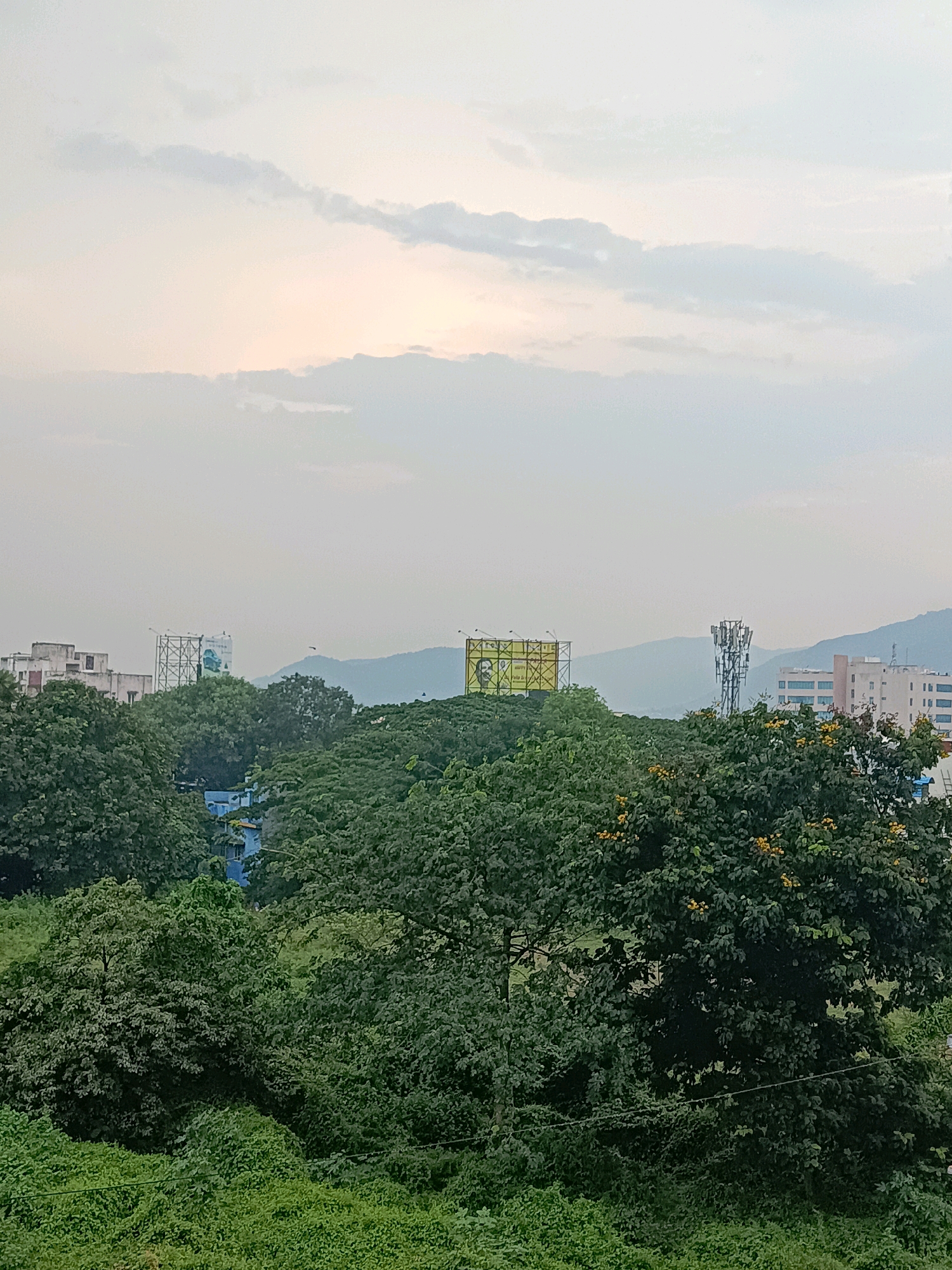 The image depicts a panoramic view of the city of Visakhapatnam, India, likely taken from an elevated position. The sky is overcast with soft sunlight peeking through the clouds, creating a diffused light across the scene. In the foreground, a dense canopy of lush green trees and vegetation dominates, with some trees bearing yellow blossoms. In the mid-ground, several buildings are visible, including a tall, multi-story structure on the left with a large billboard, and another taller building with a telecommunications tower in front of it. Further in the background, a range of hazy, blue-tinged hills forms the horizon. The overall impression is that of an urban landscape interspersed with abundant natural greenery.