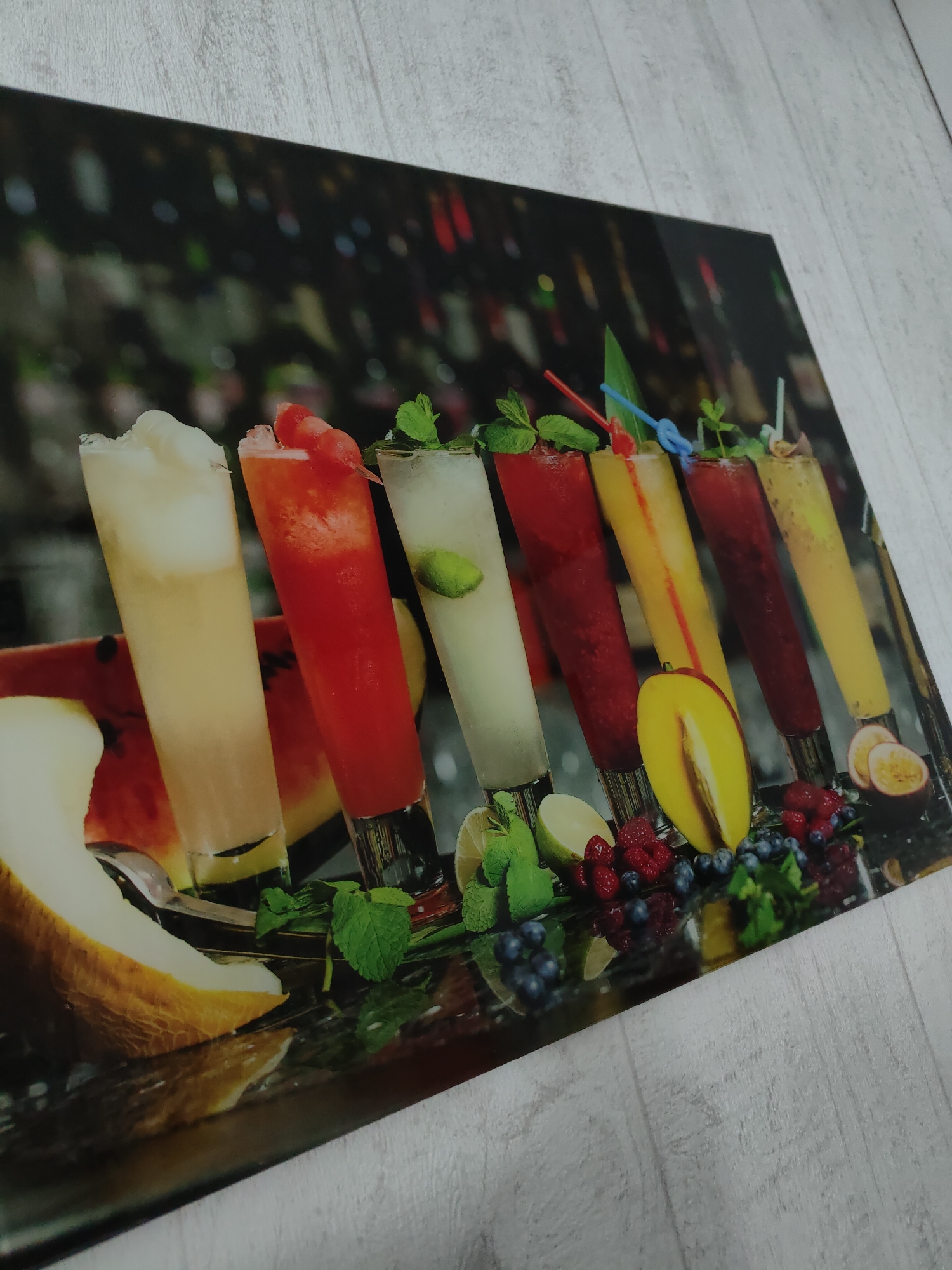 The image shows a photographic print or a picture of several tall, slender glasses filled with colorful drinks. These drinks appear to be cocktails or juices, each decorated with fresh ingredients like mint leaves, slices of fruit, and straws. Beside the glasses are various fresh fruits, including watermelon, melon, limes, blueberries, raspberries, peach, and passionfruit.
The backdrop behind the drinks is a blurred image of what seems to be a bar, with shelves containing numerous bottles of various shapes and colors.
The print is set against a light grey wooden surface. The scene does not appear to be a real location but rather a still life staged for a photograph. The image does not provide specific information related to the city of Ufa, Russia, and the context lacks any connection to this location.