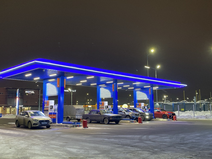 A gas station at night is illuminated by bright blue lights lining the canopy. Several cars, including a white compact, a gray sedan, a dark SUV, and a red SUV, are parked beneath it. A small trailer sits near the gas pumps. A hotel is visible in the background to the left, and streetlights shine in the distance. The ground is covered with snow.