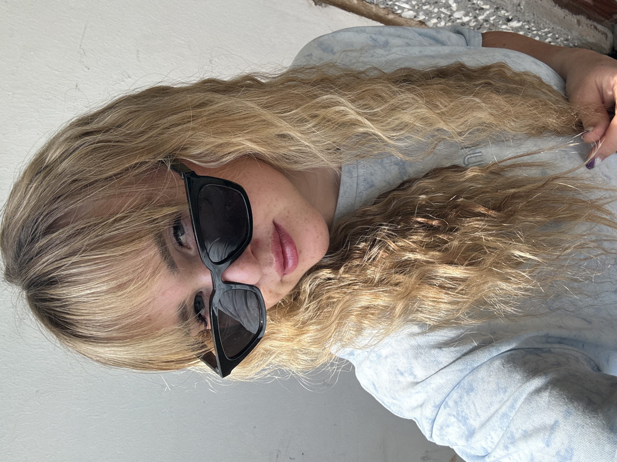 A young woman with blonde, wavy hair and bangs, wearing black sunglasses and a light blue tie-dye t-shirt, poses for a selfie indoors. She is looking towards the camera, with her mouth slightly open. The background consists of a plain white wall and some debris on the right side of the image. The lighting suggests it is daytime, possibly near a window.