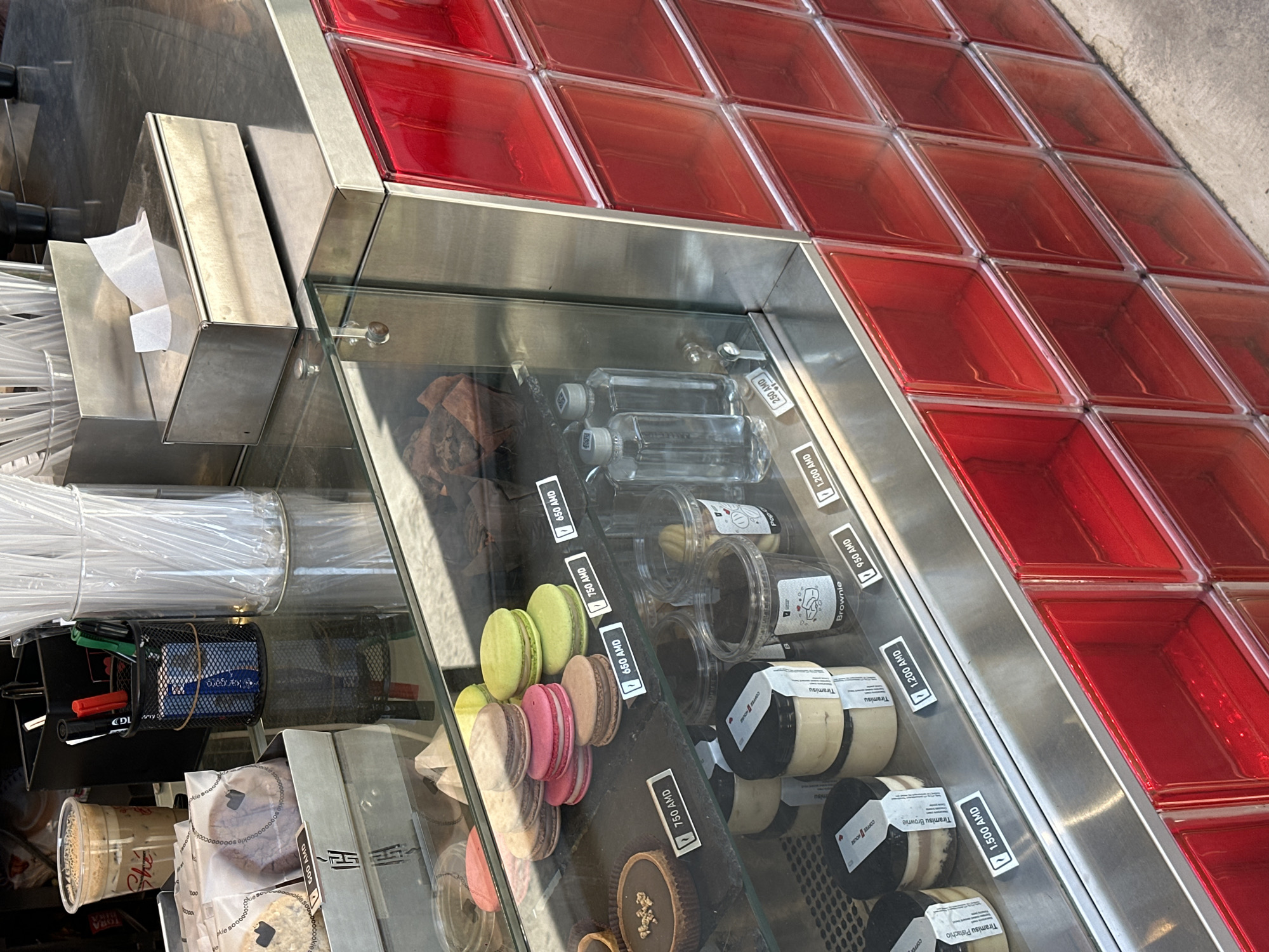 This image captures a vibrant display counter at what appears to be a cafe or dessert shop in Yerevan, Armenia. The scene is dominated by a stainless steel and glass display case, presenting an array of sweet treats and beverages.
Inside the glass display, a variety of desserts are showcased. On the left, colorful macarons in shades of light green, pink, and brown are neatly arranged, with visible price tags indicating 650 AMD and 750 AMD. Below them, round tarts, including a dark-colored chocolate one, are also displayed, some priced at 750 AMD. Various individual portions of desserts in transparent plastic jars include "Tiramisu" (1,200 AMD), "Tiramisu Brownie" (1,200 AMD), and "Tiramisu Pistachio" (1,500 AMD), many bearing "COFFEE HOUSE" branding. Small, clear plastic water bottles are priced at 250 AMD, and packaged cookies, some labeled "cookie," are available for 500 AMD. Another item, "Brownie Pop," is priced at 950 AMD.
To the left of the dessert section, a service area features a stainless steel napkin dispenser containing white napkins, a tall container filled with clear plastic straws, and a black mesh caddy holding pens.
Above and to the right of the main display case, a grid of bright red square containers is prominently featured, likely holding various ingredients or toppings for customized food or drink items. The clean lines and organization suggest an efficient and well-maintained establishment. The currency "AMD" (Armenian Dram) on all price tags firmly situates the location in Armenia, specifically Yerevan as per the provided context.