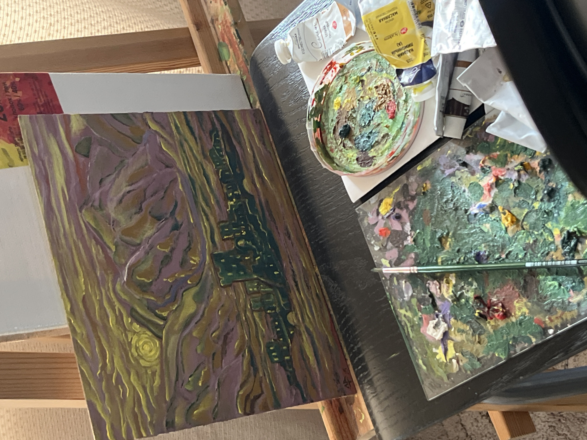 The image shows an artist's workspace. In the foreground, there's a canvas on an easel featuring an unfinished painting. The style is reminiscent of Van Gogh, with swirling brushstrokes and a color palette of yellow, purple, and green. The painting appears to depict a cityscape.
To the right of the canvas, there's a dark-colored table with painting supplies. There are tubes of paint, a round palette with a mix of colors, and a flat, rectangular palette covered in dried paint. A paintbrush rests on the rectangular palette.
The tubes of paint have labels with Russian text, including words like "Масляная Художественная" and "Кадмий лимонный". The label on the canvas has Russian text reading, "полок / лен 60%/40%".
Based on the imagery, the painting shows a landscape in Krasnoyarsk, Russia.