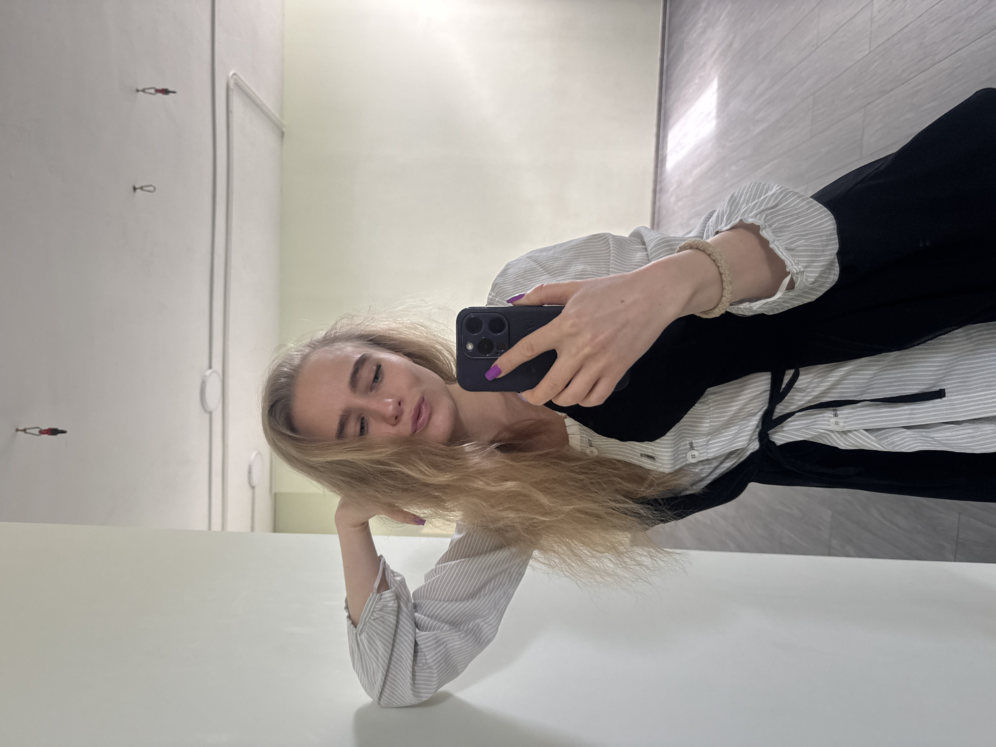The image captures a young woman taking a selfie in an indoor, brightly lit setting. She has long, wavy blonde hair and is looking towards the camera of her dark-colored smartphone, which she holds in her left hand, displaying purple nail polish. Her right arm is bent at the elbow, resting on a flat, light-colored surface, with her hand supporting her chin. She is wearing a light-colored, possibly white or light grey, striped long-sleeved shirt, with a black dress or top layered over it. A light brown, braided bracelet is visible on her right wrist. Her expression is relaxed and slightly contemplative, with her lips slightly parted.
The background consists of plain, light-colored walls. On the wall behind and to the left of her, there are a few small metallic hooks or hangers, one of which has a small red and black object attached, possibly a miniature hanger or decorative item. The floor or a vertical surface in the upper right part of the image features a subtle striped texture and a rectangular patch of bright light, suggesting a window or a strong light source. The overall lighting is bright and even, indicative of daytime. The environment is clean and minimalist, potentially a changing room, a modern studio, or a simple room in a home. There is no visible text in the image. Location context is Unknown.