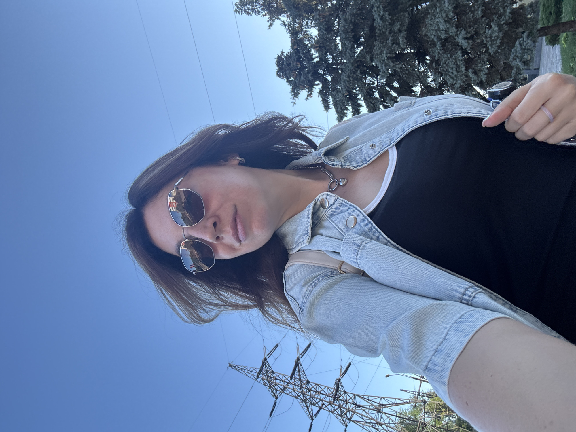 A woman with long brown hair and sunglasses is standing outdoors in front of a clear blue sky, with a large metal power pylon to her left and evergreen trees to her right. She is wearing a light blue denim jacket over a black top, a silver necklace with a heart-shaped pendant, and a light pink ring on her right hand. The sunglasses reflect buildings and roads. The location appears to be Kryvyi Rih, Ukraine, likely in an urban or industrial area with power infrastructure.