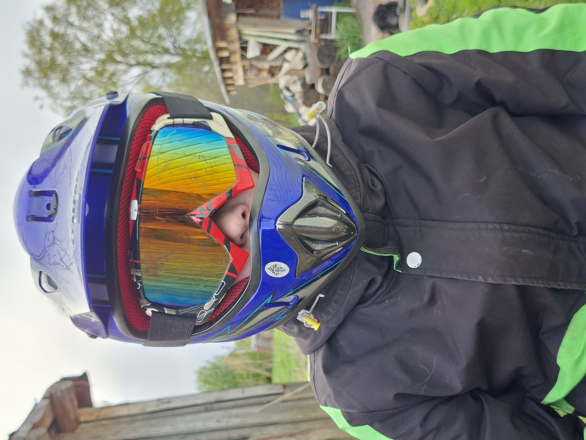 A close-up shot shows a person wearing a blue and black motocross helmet with goggles, reflecting a vibrant, distorted view of the surrounding environment. The person is wearing a black jacket with green trim. The background is slightly blurred, but appears to be an outdoor setting with a building and some foliage visible. The exact location in the city of Unknown, Unknown, cannot be determined from the provided image.