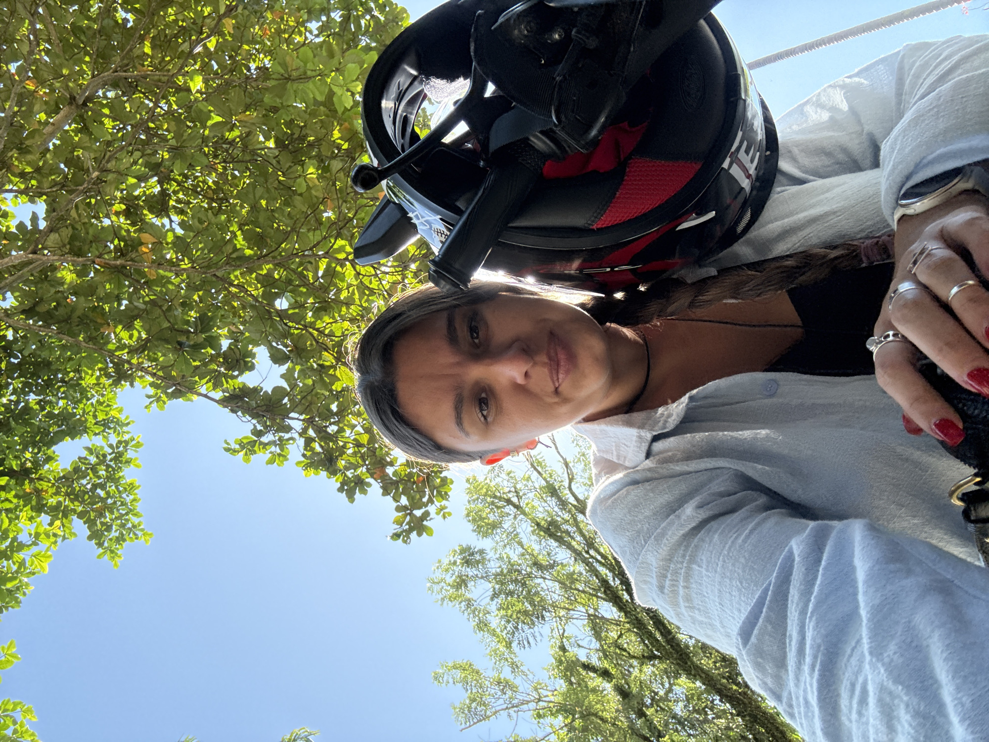 A ground-level, upward-angled selfie shows a woman wearing a black and red motorcycle helmet and a light blue long-sleeved button-down shirt. She is looking directly at the camera with a neutral expression. A black handbag strap is visible near her hand, which is adorned with multiple silver rings and has red nail polish. Above and around her, green foliage from trees fills much of the background, contrasting with patches of clear blue sky. Based on the foliage and the general ambiance, the location is likely in a natural or semi-rural setting within the Ubud District, Indonesia.