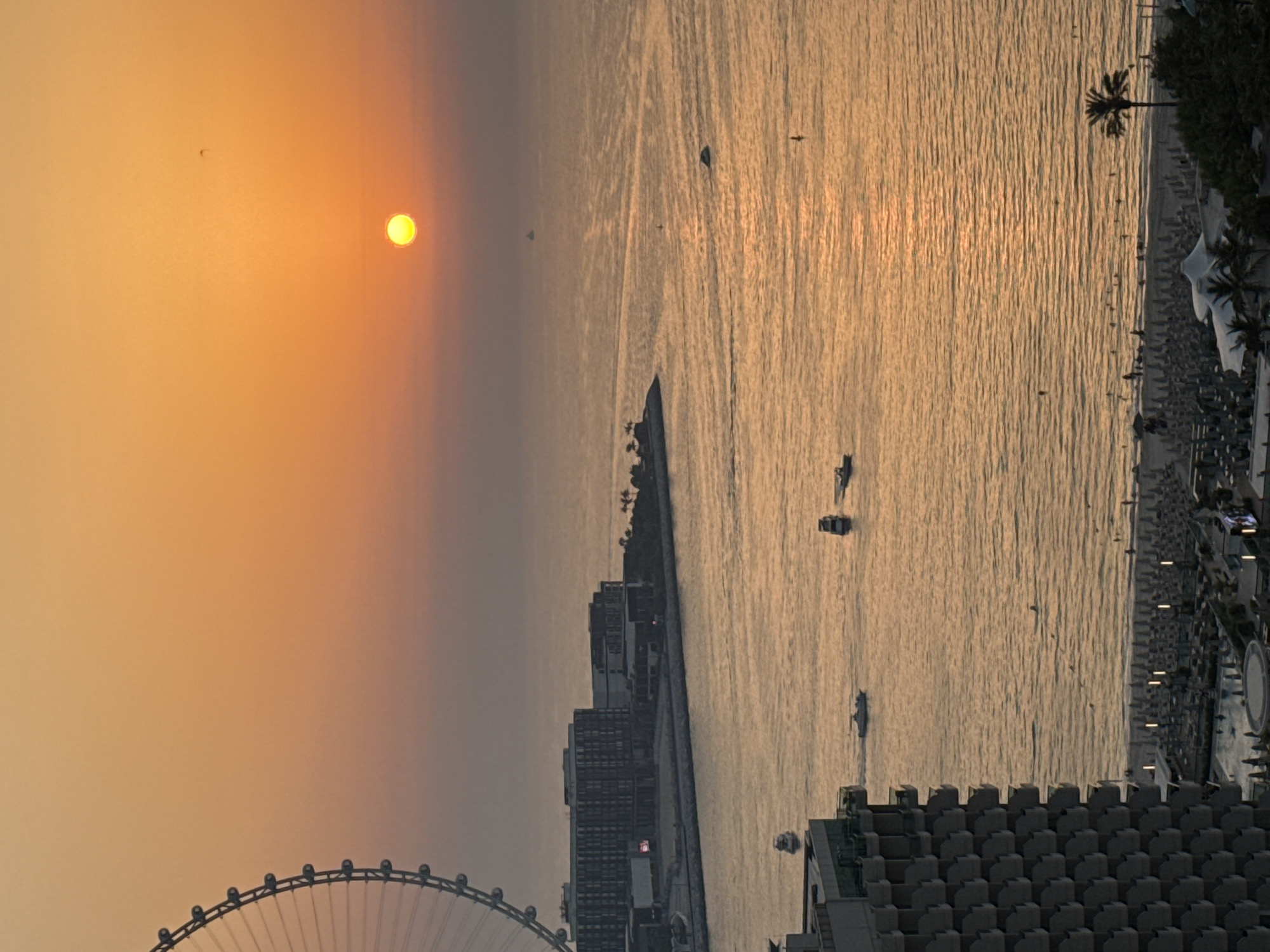 This is a vertical, eye-level photograph capturing a sunset over a city waterfront, likely in Dubai, UAE. The sun, a bright yellow orb, is low on the horizon, casting a warm, orange glow across the sky and water. The water's surface is textured with ripples, reflecting the golden light and creating shimmering patterns.
Several boats of varying sizes are visible on the water. In the foreground on the left, a portion of a large Ferris wheel is visible, its spokes extending outwards. On the right side of the image, a coastline is lined with buildings, palm trees, and what appears to be a beach with numerous lounge chairs. The buildings are modern, with numerous windows, and some have lights on. A few scattered figures can be seen on the beach and along the waterfront. The overall atmosphere is serene and tranquil, with the setting sun bathing the scene in a peaceful, warm light. The time of day is clearly sunset, and the weather appears to be clear. There is no visible text in the image.