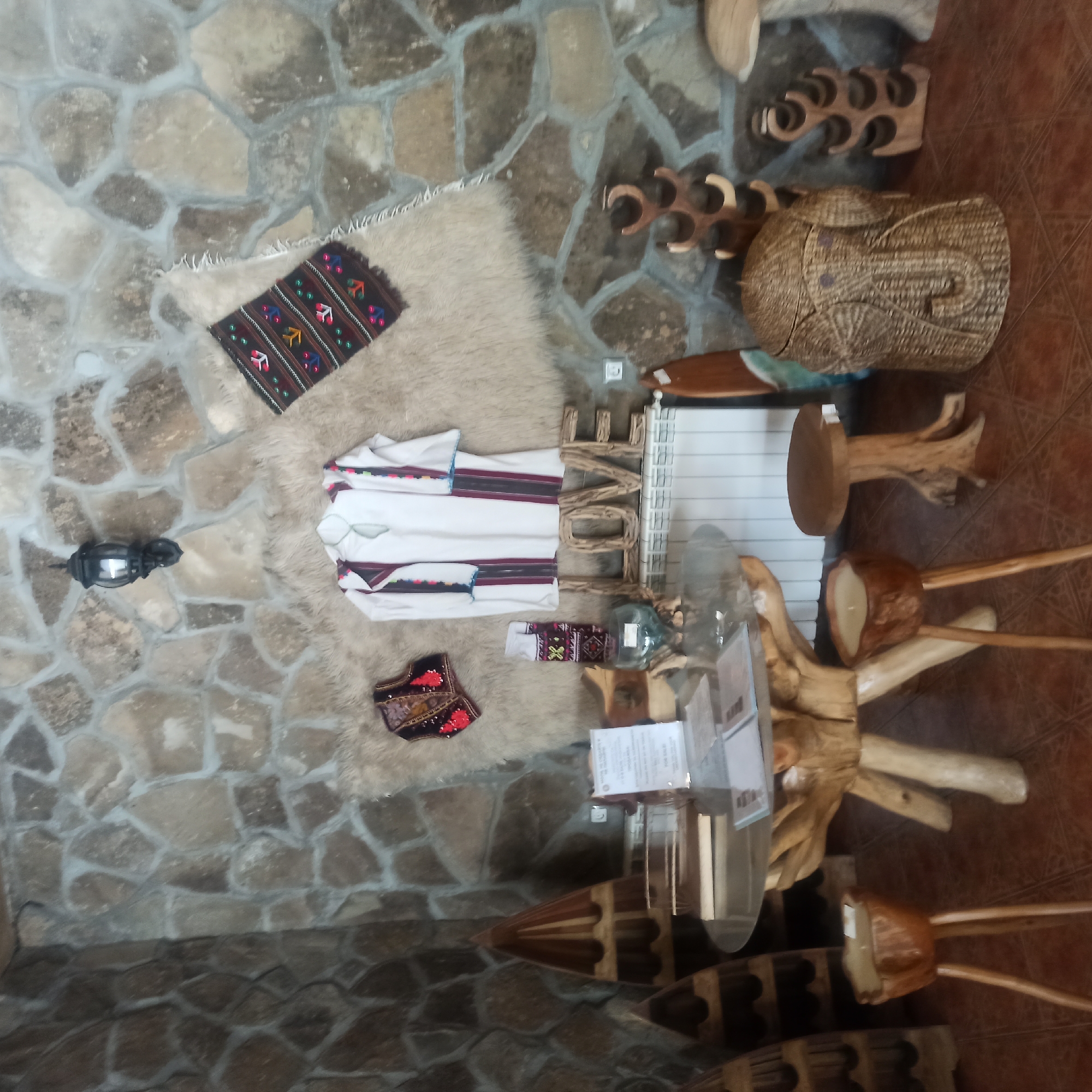 The image depicts an indoor scene in Zlatna Panega, Bulgaria, likely a gift shop or cultural center, featuring rustic and handcrafted items. The walls are constructed of irregular natural stones, creating a rugged texture. A traditional embroidered shirt and vest, along with a patterned textile, are displayed against a shaggy, cream-colored rug. The word "LOVE" is spelled out in large wooden letters on a white radiator. A round glass-topped table with a unique wooden base is in the foreground, surrounded by small, carved wooden stools and a decorative wooden planter with a candle. To the right, a woven basket shaped like an elephant stands near wooden wine racks. The overall ambiance suggests a focus on folk art and natural materials. There are no people visible in the image.
