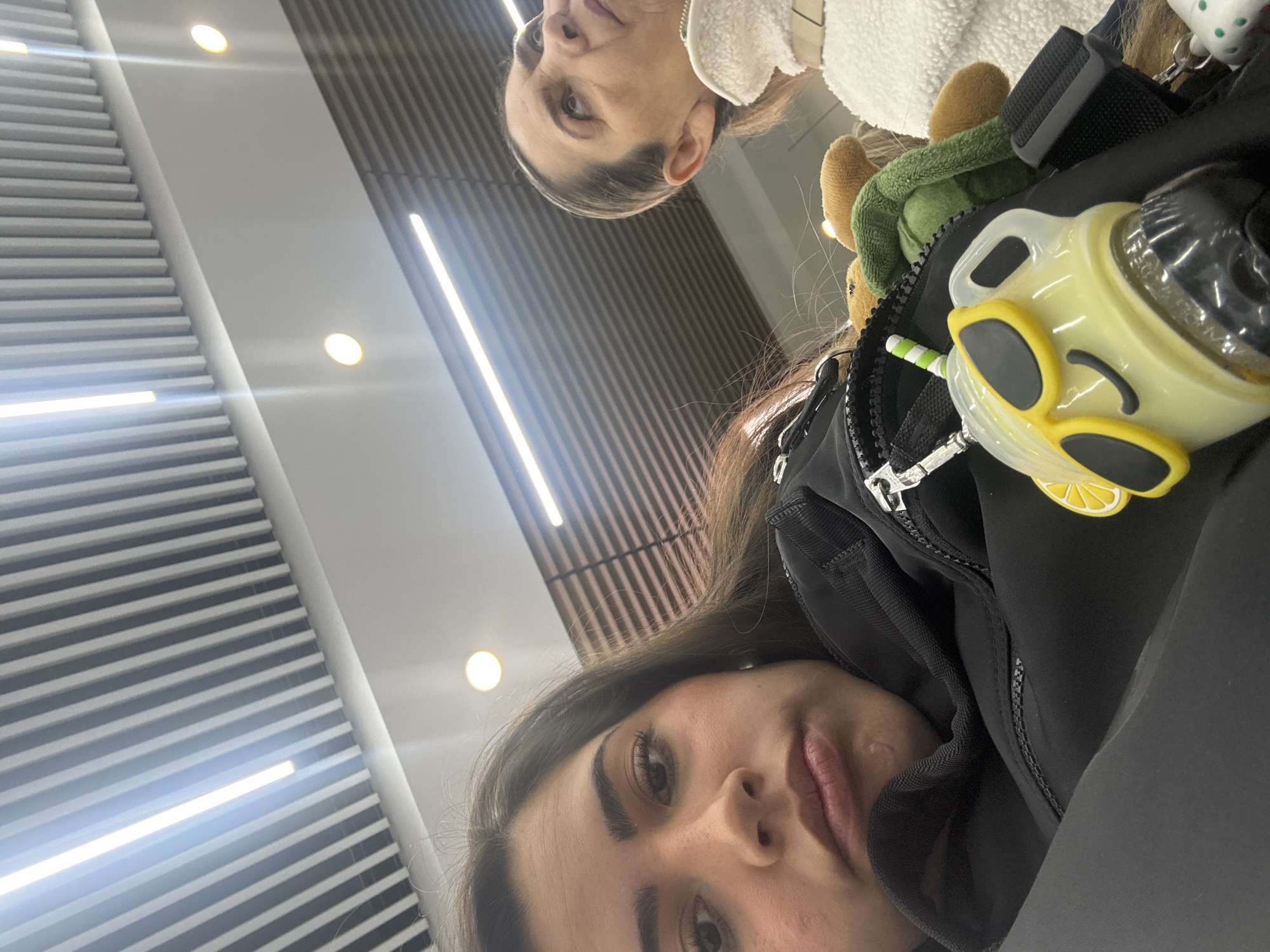 Two people are in the image. One person, with brown hair, is taking a selfie. A person with fair hair is to the upper right. The background suggests a waiting area with horizontal lines on the wall and ceiling and rows of fluorescent lights. The location is potentially in Mexico City, Mexico. The person taking the selfie has a black jacket with a lemon-themed ornament, and a plush turtle is attached.