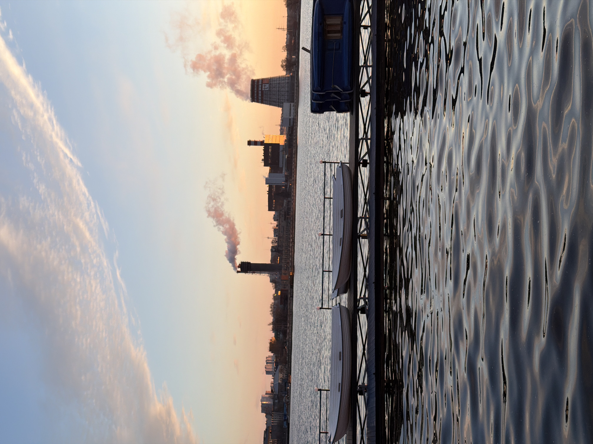 This media file captures a tranquil yet industrial scene at either sunrise or sunset, looking across a body of water towards a cityscape. The image has a horizontal orientation if viewed upright.
In the foreground, a calm expanse of water, likely a river or lake, stretches across the bottom portion of the frame. Its surface is covered with gentle ripples, reflecting the ambient light. On the right side, a dark-colored pier or dock extends into the water, where three small boats are moored. One boat, closest to the foreground, is blue, while the other two, further along the pier, are white.
Across the water, on the far bank, an industrial landscape dominates the middle ground. Prominent features include a large, grey, cooling tower-like structure with the Cyrillic letters "ИЖ" (likely an abbreviation for Izhevsk) clearly visible on its side. Several other factory buildings and smokestacks are scattered along the bank, with plumes of smoke or steam rising from at least two chimneys, tinted orange by the low sun. Beyond the industrial complex, the silhouettes of other city buildings can be seen.
The background is filled with a dynamic sky, characteristic of either dawn or dusk. It transitions from a soft blue on the left to warm hues of orange, pink, and yellow on the right, suggesting the sun is low on the horizon. Scattered, elongated clouds drift across the sky, catching the warm light.
The scene combines natural beauty with human development, depicting an early morning or late evening in Izhevsk, Russia, with no direct human activity visible.