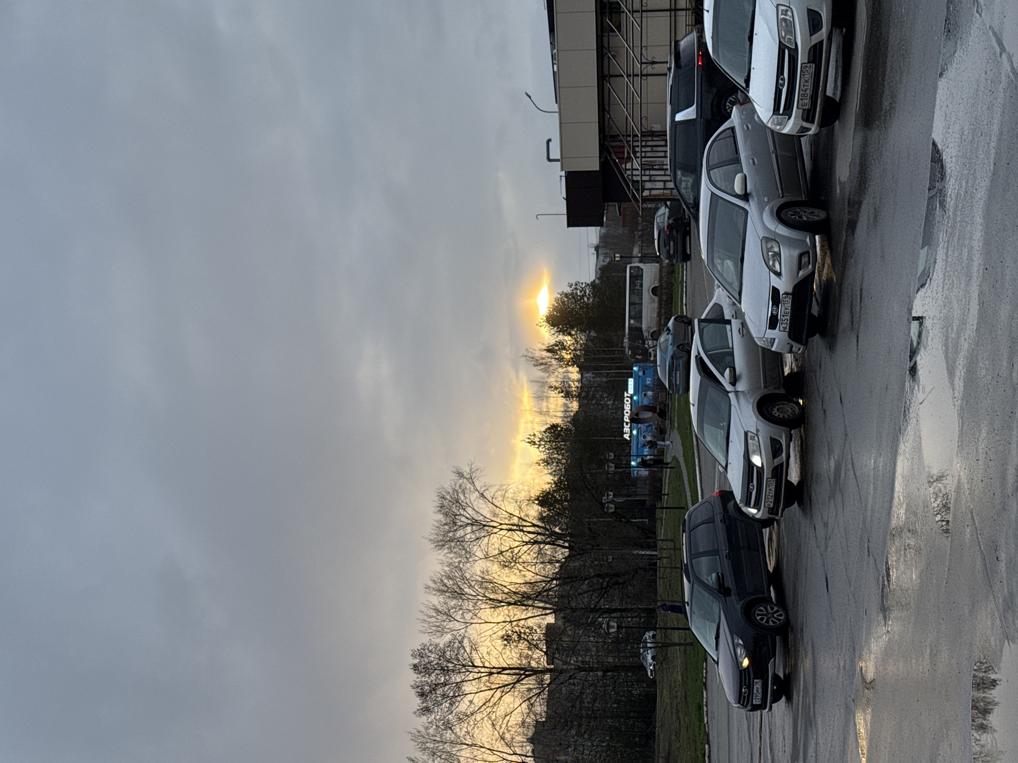 The image, when viewed upright, captures an urban or suburban scene during either sunrise or sunset, with a low sun breaking through an overcast sky. The sky is predominantly gray and white, with a bright, warm glow of orange and yellow emanating from the sun near the horizon on the left.
In the foreground and mid-ground, a row of vehicles is parked alongside a wet asphalt road or parking lot on the right side of the frame. The road surface is dark and highly reflective, indicating recent rain and featuring visible puddles that mirror the sky and cars. Prominent vehicles include several white sedans, a silver sedan, and a dark SUV. Two clear Russian license plates are visible: "E184TK 159" on a white car and "M351EM 159" on a silver car, both signifying the Perm Krai region.
On the right side, behind the parked cars, a portion of a modern building with a multi-story structure and some decorative metalwork is visible. To the left, a line of bare deciduous trees forms a dark silhouette against the brightly lit horizon. In the distance, a blue sign with white Cyrillic text "АВС РОБОТ" (ABC ROBOT) is discernible. Other vehicles, possibly buses, are faintly visible further back. No distinct people are present in the scene. The overall impression is quiet and atmospheric, marked by the contrast between the warm sunlight and the cool, damp environment.