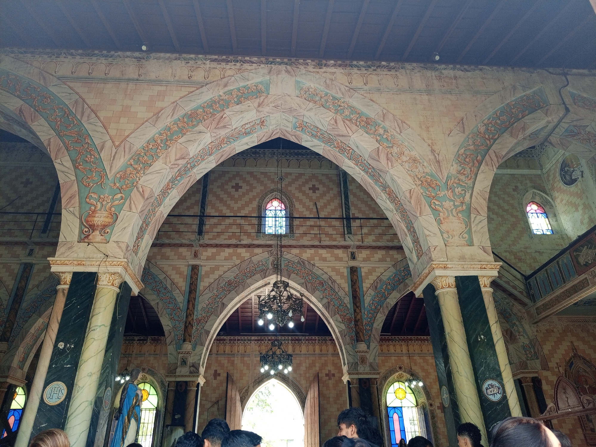 The image captures the interior of a church with a series of arches. The arches are decorated with floral designs and the columns supporting them appear to be made of marble or a similar stone. Chandeliers hang from the ceiling, which is made of wood planks. Stained glass windows can be seen in the background. There are people present in the lower portion of the image, suggesting a service or event is taking place. The image is taken inside the church in Tinaquillo, Venezuela.