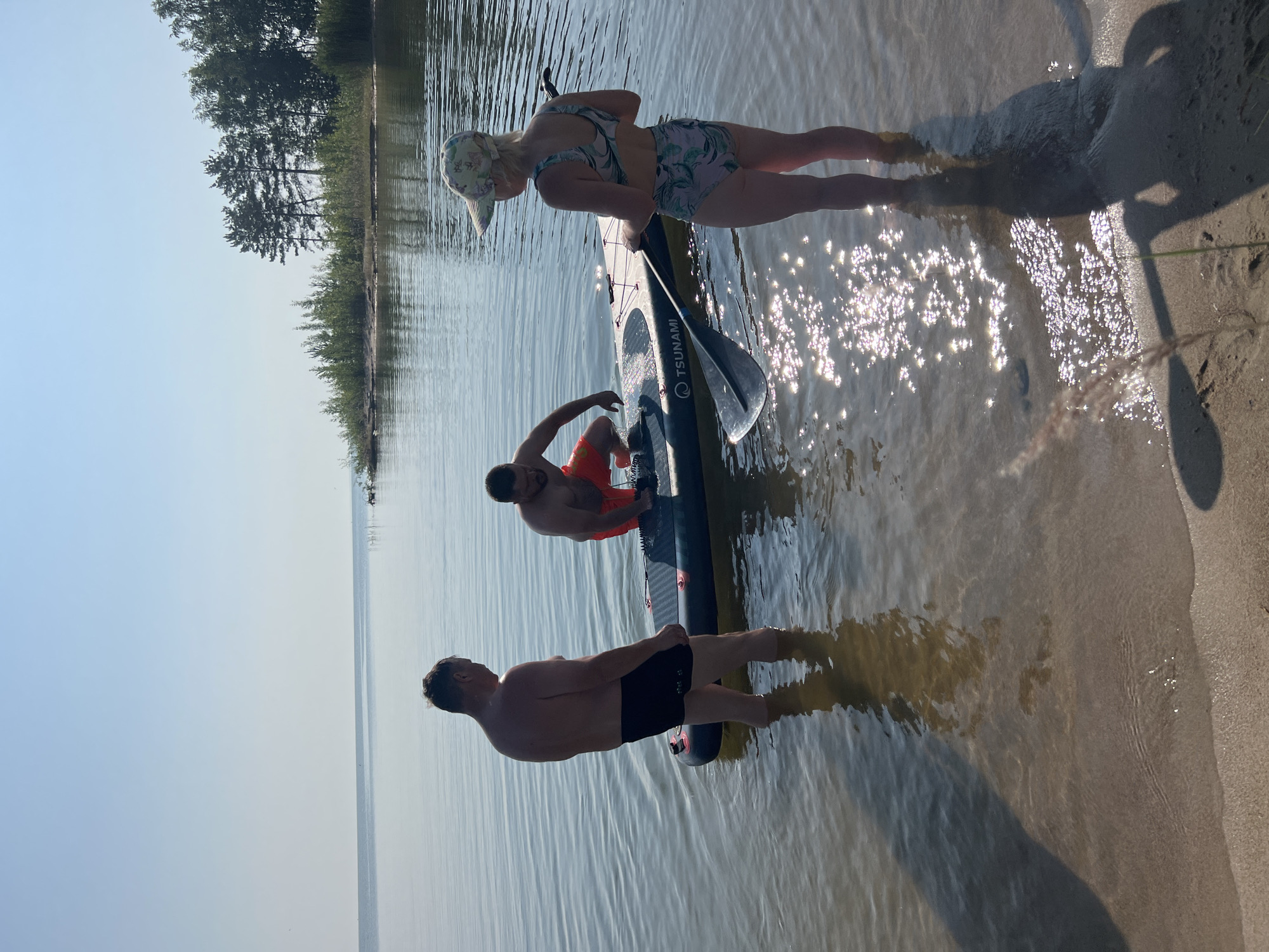 Three individuals are in shallow, calm water near the shore of a large body of water, likely a lake, under a clear blue sky. A paddleboard is floating in the water between them. One man, wearing black swim trunks, stands facing away from the camera. Another man, in orange swim trunks, is crouched on the paddleboard, appearing to adjust something. A woman, wearing a patterned bikini and a cap, stands facing the paddleboard and the other two individuals, holding a paddle. The environment suggests a sunny day at a natural recreation area, with trees visible along the distant shoreline. The water is clear enough to see the sandy bottom. The image captures a moment of preparation or activity related to the paddleboard.