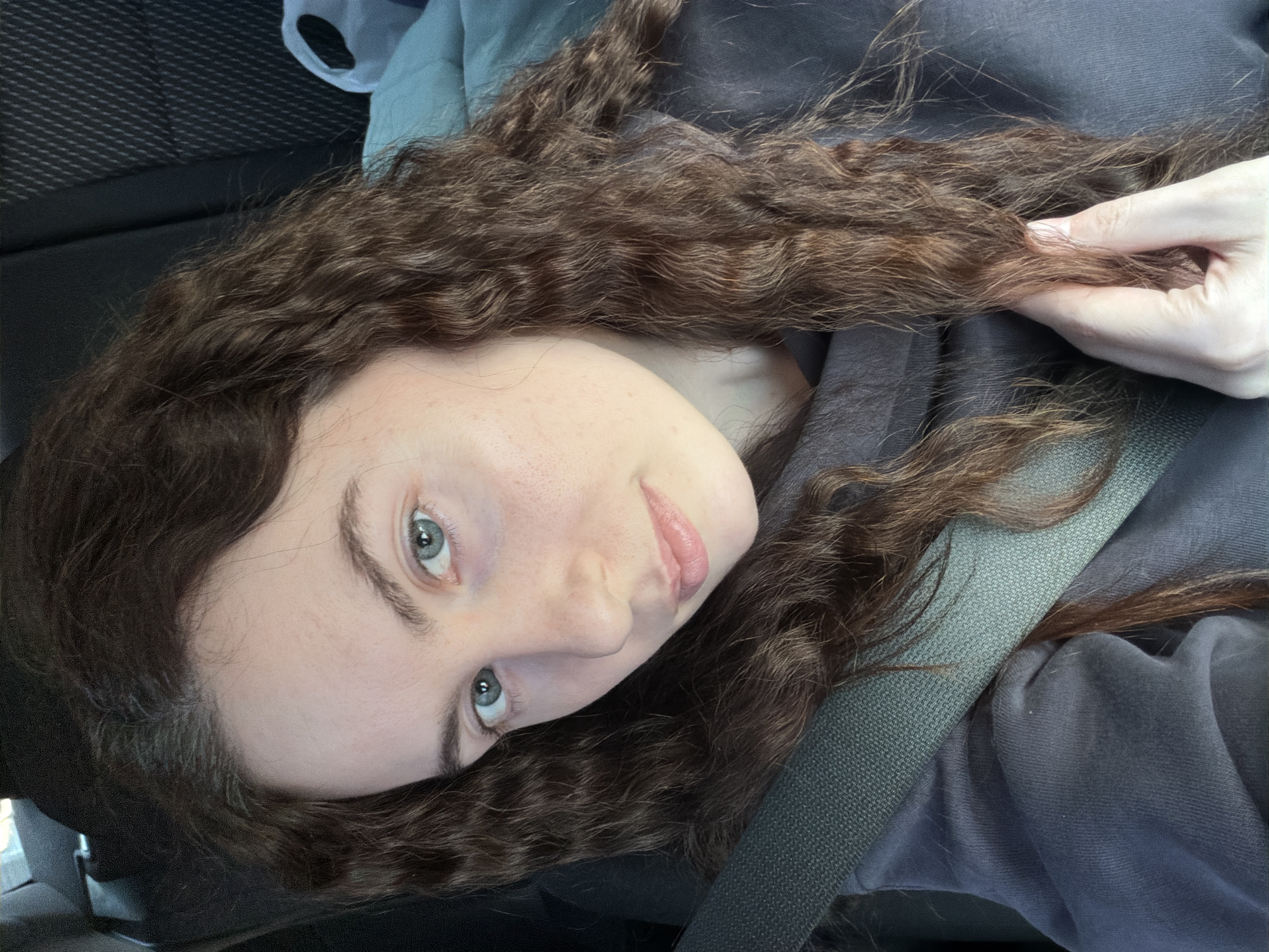 A young woman with light skin, brown curly hair, and light blue eyes is wearing a dark grey hooded sweatshirt and a dark grey seatbelt. She is holding a strand of her hair in her right hand. She is located in a vehicle. The location is not evident from the image and it cannot be determined if the scene is in the city of Saint Petersburg, Russia.