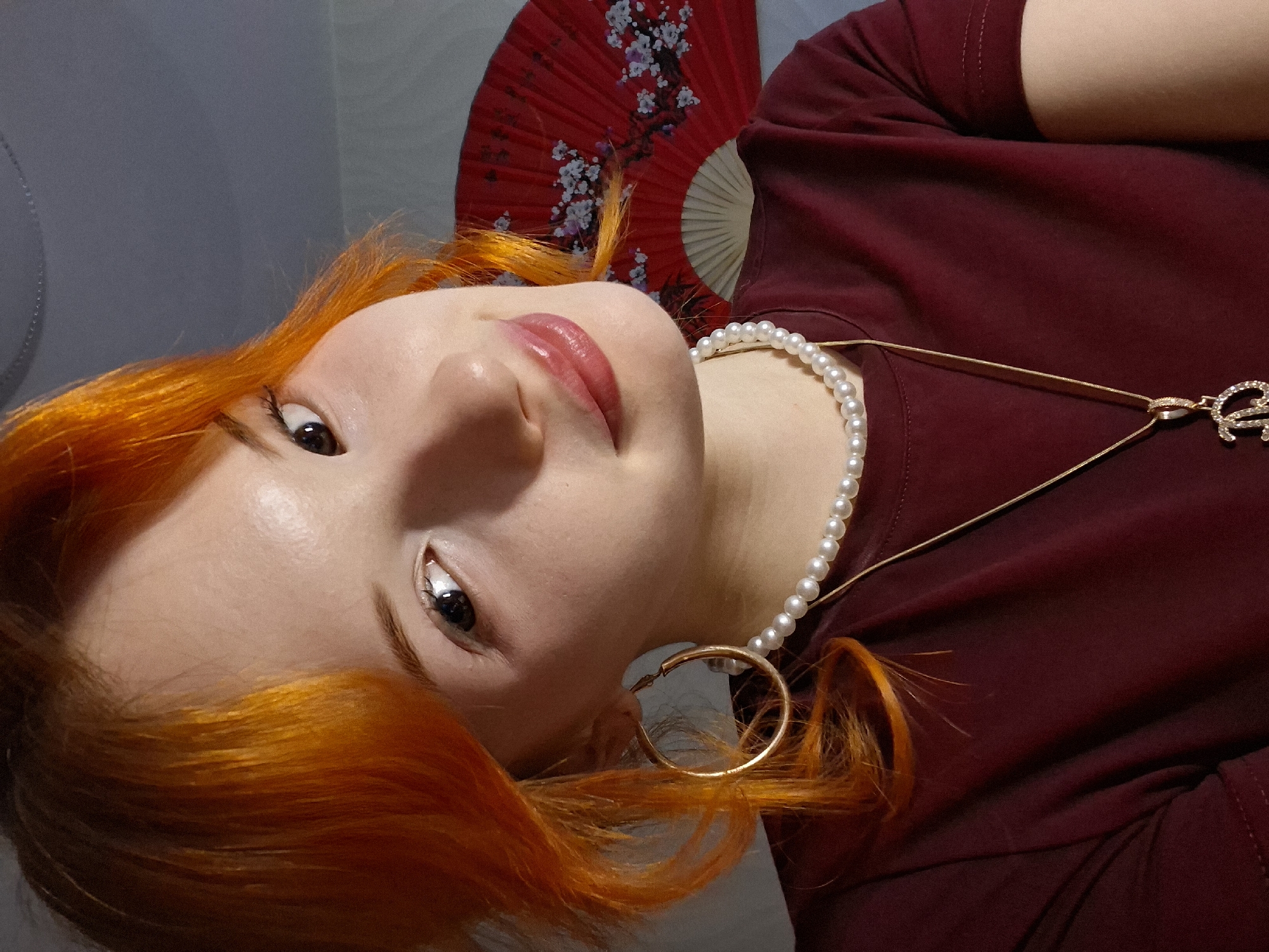 A close-up, slightly angled, indoor portrait features a fair-skinned individual with bright orange hair. She is wearing a maroon-colored top and a pearl necklace. A gold-colored necklace with a pendant is also visible. Her lips are glossy with a reddish hue, and she has dark eyes. A gold hoop earring is visible on one side of her face. A red hand fan with white floral designs is partially visible in the background.
There is no information available in the image to indicate that this portrait was taken in Chaikovsky, Russia.