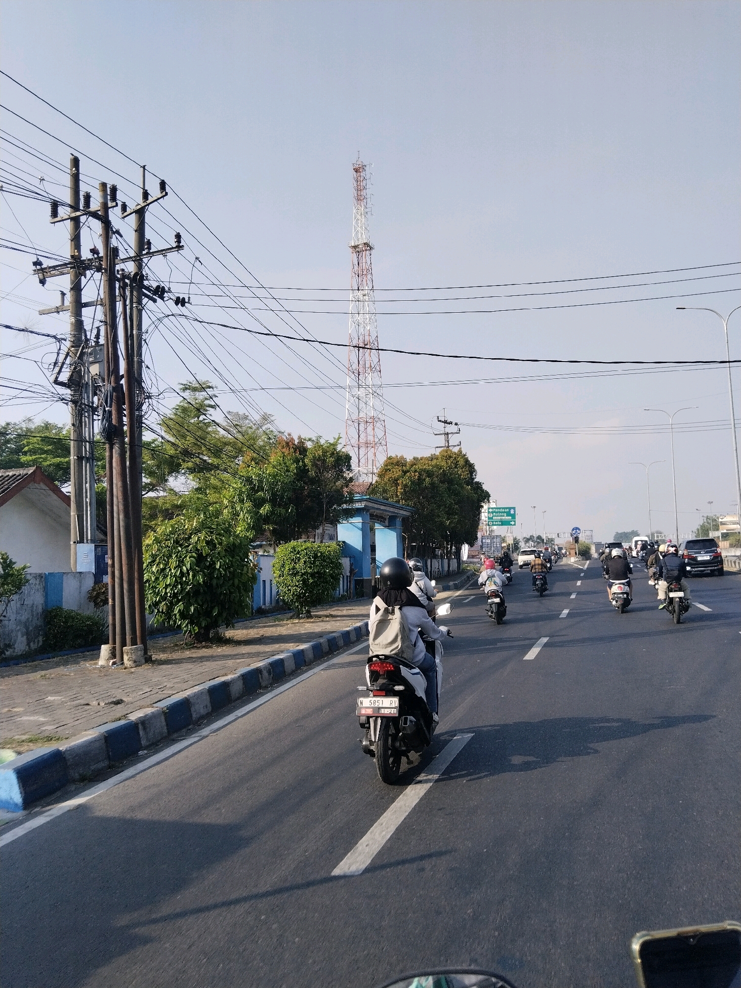 This image depicts a street scene in Banjararum, Indonesia, with a focus on a busy road and its surroundings. The road is paved and marked with white lines, carrying a moderate amount of traffic, primarily motorcycles. Several individuals are visible riding motorcycles, some with passengers. They are wearing helmets for safety. In the background, a tall, red and white telecommunications tower stands prominently against a clear, pale blue sky. To the left of the road, there are power lines supported by utility poles and trees lining the sidewalk. A building with a tiled roof is also visible on the left. On the right side of the road, more vehicles, including cars and motorcycles, are traveling. Streetlights are present, indicating it's an urban or semi-urban area. The overall atmosphere appears to be a typical day in a moderately busy Indonesian town.