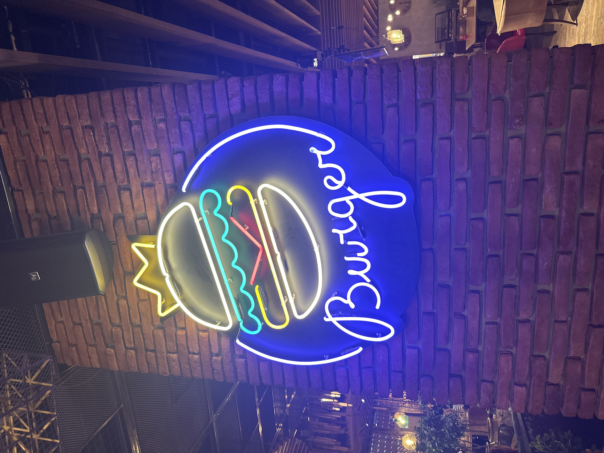 The image captures a neon sign shaped like a burger with the word "Burger" written below it in a script font. The neon lights are a vibrant blue, white, yellow, and red. The sign is mounted on a brick wall in what appears to be an indoor establishment, possibly a restaurant or bar. Above the sign, a black speaker with an "EV" logo is visible. The background shows wooden beams and a ceiling structure, suggesting an industrial or rustic interior design. In the lower right portion of the image, tables and chairs are visible, indicating a dining area. There are no people clearly depicted in the scene. The lighting in the image is dim and cast with a purple hue, likely from the neon sign and ambient lighting. This scene is located in the city of Varna, Bulgaria.