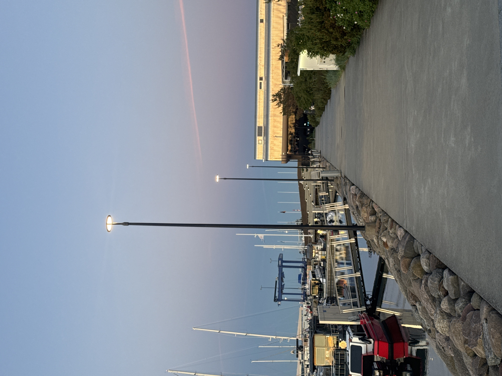 A waterfront scene in Tallinn, Estonia, captures a harbor with several boats docked. A paved walkway lines the harbor, bordered by large rocks on one side and a building and vegetation on the other. Lampposts are spaced along the walkway, illuminating the scene. In the background, sailboats and a large blue crane are visible, suggesting a working harbor. The sky displays a soft gradient of blue and pink, indicating either sunrise or sunset. A red and white boat is prominently featured in the foreground, reflecting in the calm water. The overall atmosphere is serene and peaceful.