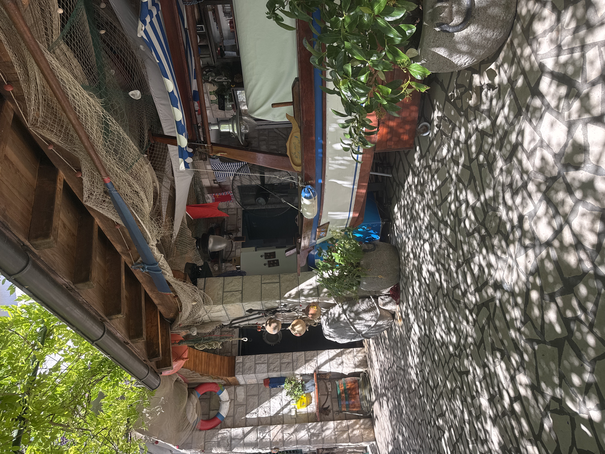 The image displays an outdoor, narrow passage or courtyard adorned with a strong nautical theme, viewed from a slightly high angle. No people are present.
The central subject is a wooden boat, possibly a display piece, with white, blue, and brown hull details, positioned to the right of the frame. It features a visible rudder or tiller, a mast, and what appears to be a light green sail partially furled. A black electric fan is visible beneath the boat.
The scene is heavily decorated with maritime items: fishing nets are extensively draped across the upper left side, over wooden beams, and near the boat, some adorned with small shells or floats. Several orange, white, and red buoys, including a prominent red, white, and blue life preserver, are visible. Hanging from a string are smaller decorative floats. Wooden oars or paddles lean against structures.
The ground is paved with irregular light-colored stone tiles, creating a mosaic pattern. Bright sunlight casts strong, dappled shadows across the entire ground, indicating a clear, sunny day. The shadows suggest overhead foliage or intricate structures.
On the left, an open wooden structure with a dark rain gutter runs along the top, beneath which is green foliage, possibly from an overgrown tree or bush. On the right, large stone planters hold green leafy plants, some with small berries or flowers. The background features light-colored stone walls, with what might be a dark door or entrance featuring a porthole-like window in the distance. A wooden barrel is also visible. No discernible text is visible in the image.