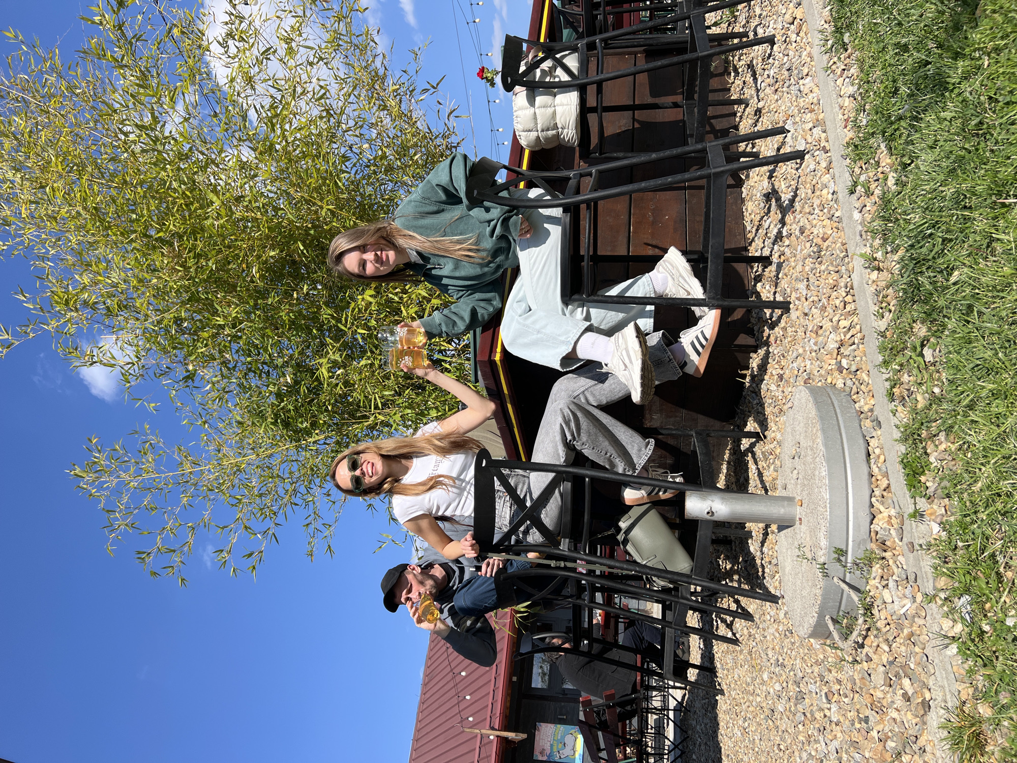 This media file captures three individuals enjoying drinks outdoors on what appears to be a sunny day in Belgrade, Serbia. The image is oriented sideways, with subjects seated at a high-top table.
Two women and one man are the primary subjects. The woman on the left (when viewed correctly) is wearing a dark green hoodie, light-colored pants, and white sneakers with dark stripes. She holds a glass filled with an amber liquid. Next to her, another woman with long blonde hair and sunglasses on her head wears a white t-shirt and grey jeans. She is also holding a glass of amber liquid, and her other arm is raised in a gesture of celebration or greeting. The man on the right is wearing a dark baseball cap and a dark jacket, holding a glass of the same amber liquid. All three are smiling, indicating a cheerful and relaxed atmosphere.
They are seated on tall black metal chairs around a table. Behind them, a dark wooden structure is visible, likely part of the seating area. A red rose in a small pot is placed on a ledge. String lights are strung overhead, and a lush green plant with bamboo-like leaves frames the left side of the scene. The ground surface features small gravel bordered by a strip of concrete and green grass. A large, circular metal base, possibly for an umbrella stand, is visible on the gravel. In the background, part of a building with a reddish roof is discernible.
The weather is bright and sunny, with a clear blue sky and a few white clouds, suggesting it is daytime. Shadows are cast by the chairs and people, confirming strong sunlight. The overall mood is one of enjoyment and social gathering on an outdoor patio or beer garden. There is some partially visible, unclear text on the white t-shirt worn by the woman in the middle.