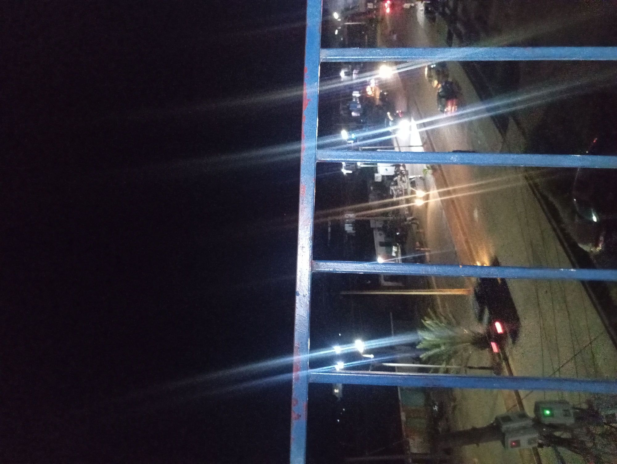A low-angle night shot captures a street scene through a blue metal railing. The sky is a deep, starless black, with bright light streaks emanating from streetlights and vehicle headlights. In the foreground, the blue railing, showing signs of rust on its upper edge, partially obstructs the view of the street below.
The street is paved and appears to be a two-lane road, with several cars visible. One car, with its brake lights illuminated, is parked on the right side of the road. Another car is driving away from the camera, its taillights glowing red. Further down the street, streetlights cast a warm glow on the asphalt.
On the left side of the street, a traffic signal box with a green light is visible, along with a palm tree. Buildings and structures are dimly lit in the background. The overall atmosphere is that of a quiet urban street at night.