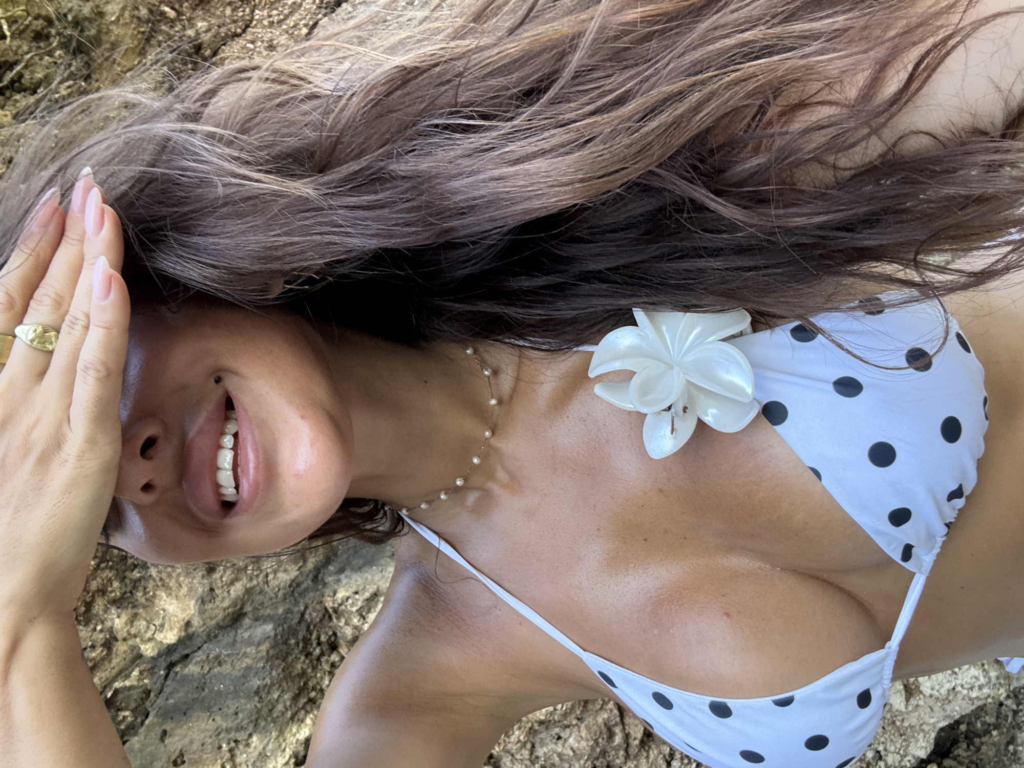 A woman is lying on a rocky surface, possibly in Kuta Selatan, Indonesia. She is wearing a white bikini with black polka dots and a white flower. She is also wearing a pearl necklace and a ring on her finger. Her hair is long and brown. She is smiling and covering her face with her hand.