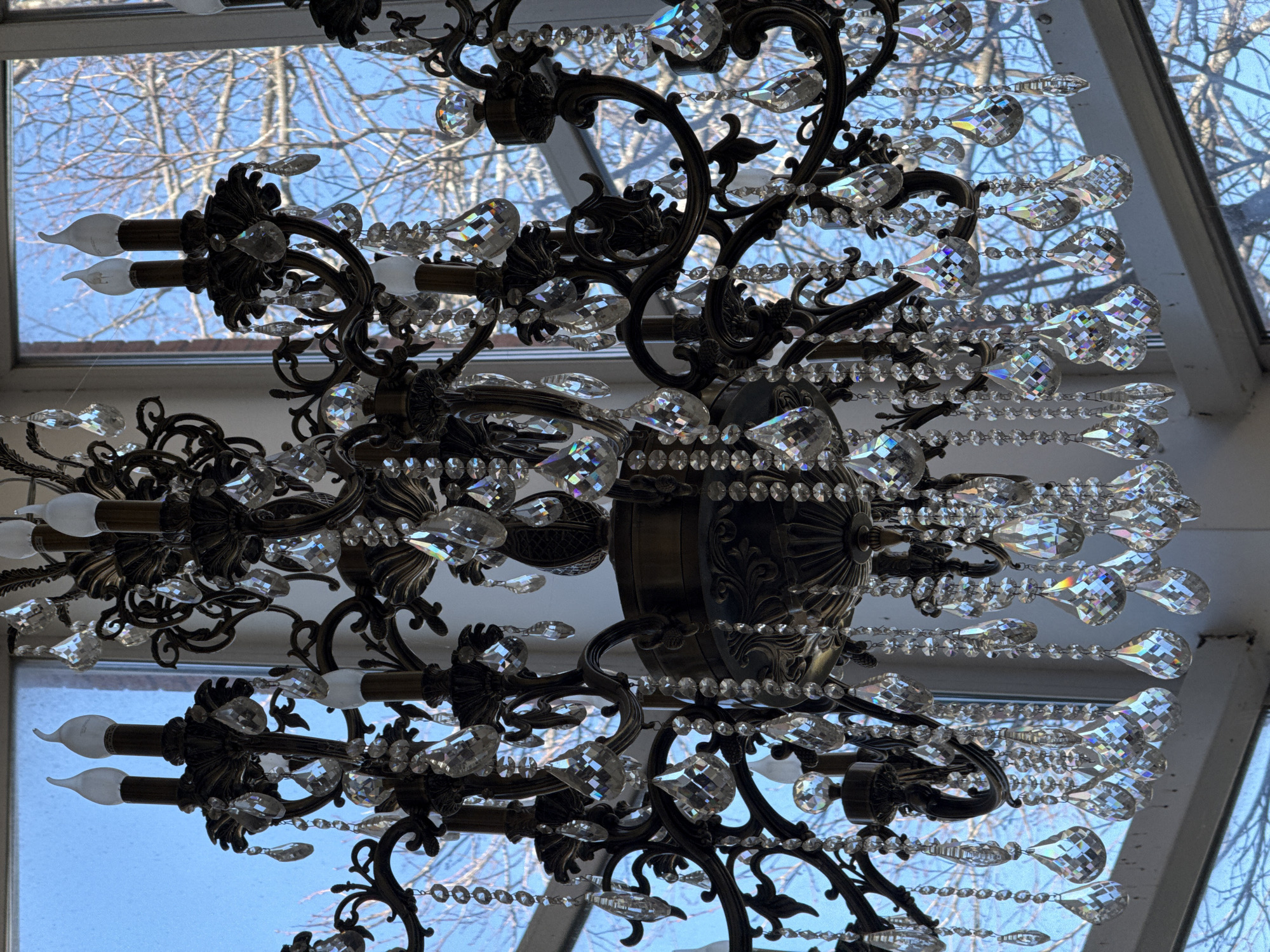 A detailed, low-angle shot captures a chandelier with an elaborate, dark metal frame adorned with numerous clear, sparkling crystals. The chandelier features several candle-style bulbs, emitting a soft light. In the background, through what appears to be a window or skylight, bare tree branches are silhouetted against a pale blue sky. The location is Galevo, Russia.