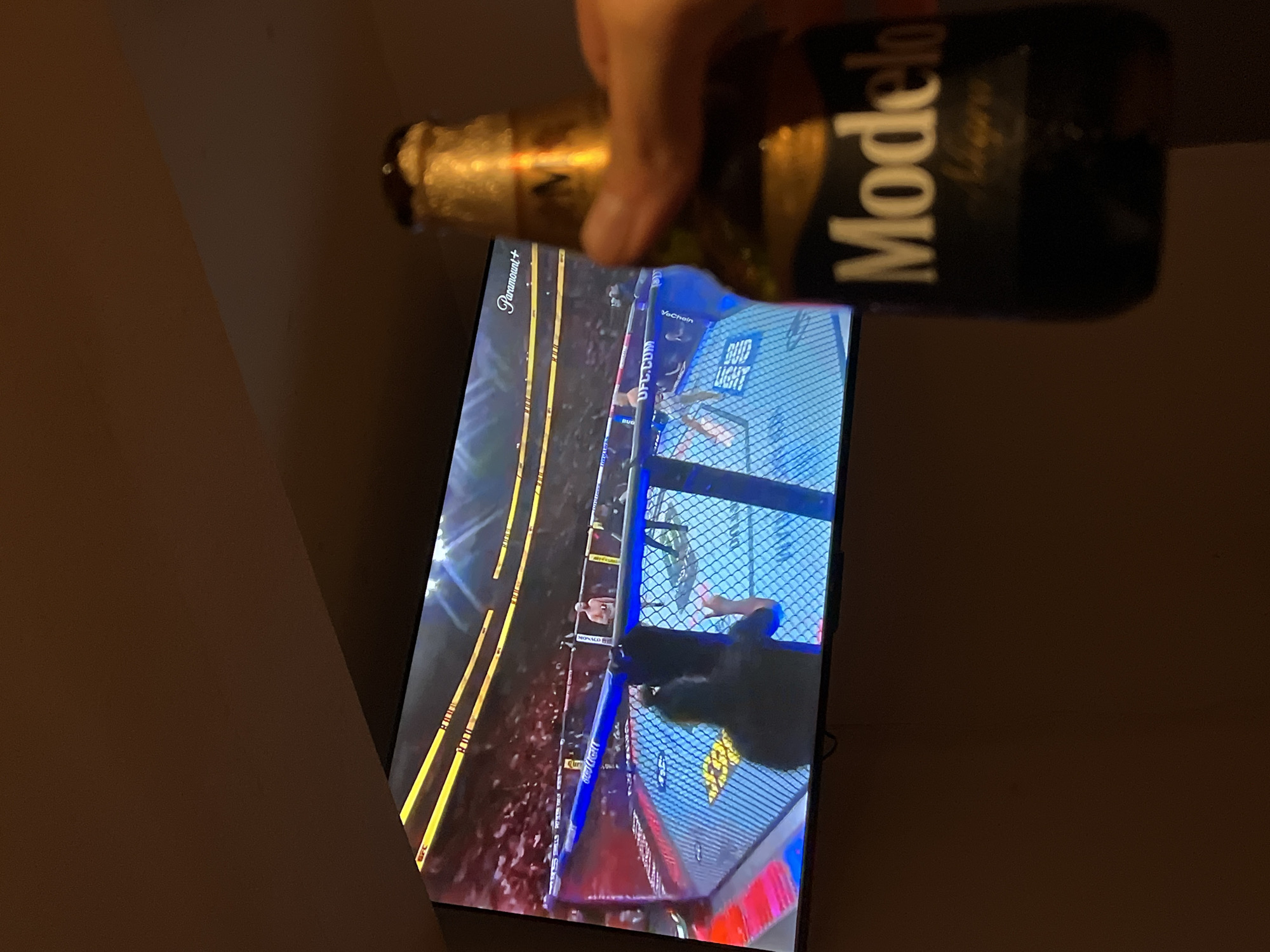 The image shows a Modelo beer bottle being held in front of a television screen displaying a UFC fight. The screen is displaying a mixed martial arts cage fight, sponsored by Bud Light and VeChain, and streaming on Paramount+. The location is not evident in the image and therefore I cannot provide a concise, forensic, matter-of-fact description of the scene, including essential details about people, subjects, objects, actions, interactions, and the environment in San Bernardino Tlaxcalancingo, Mexico.