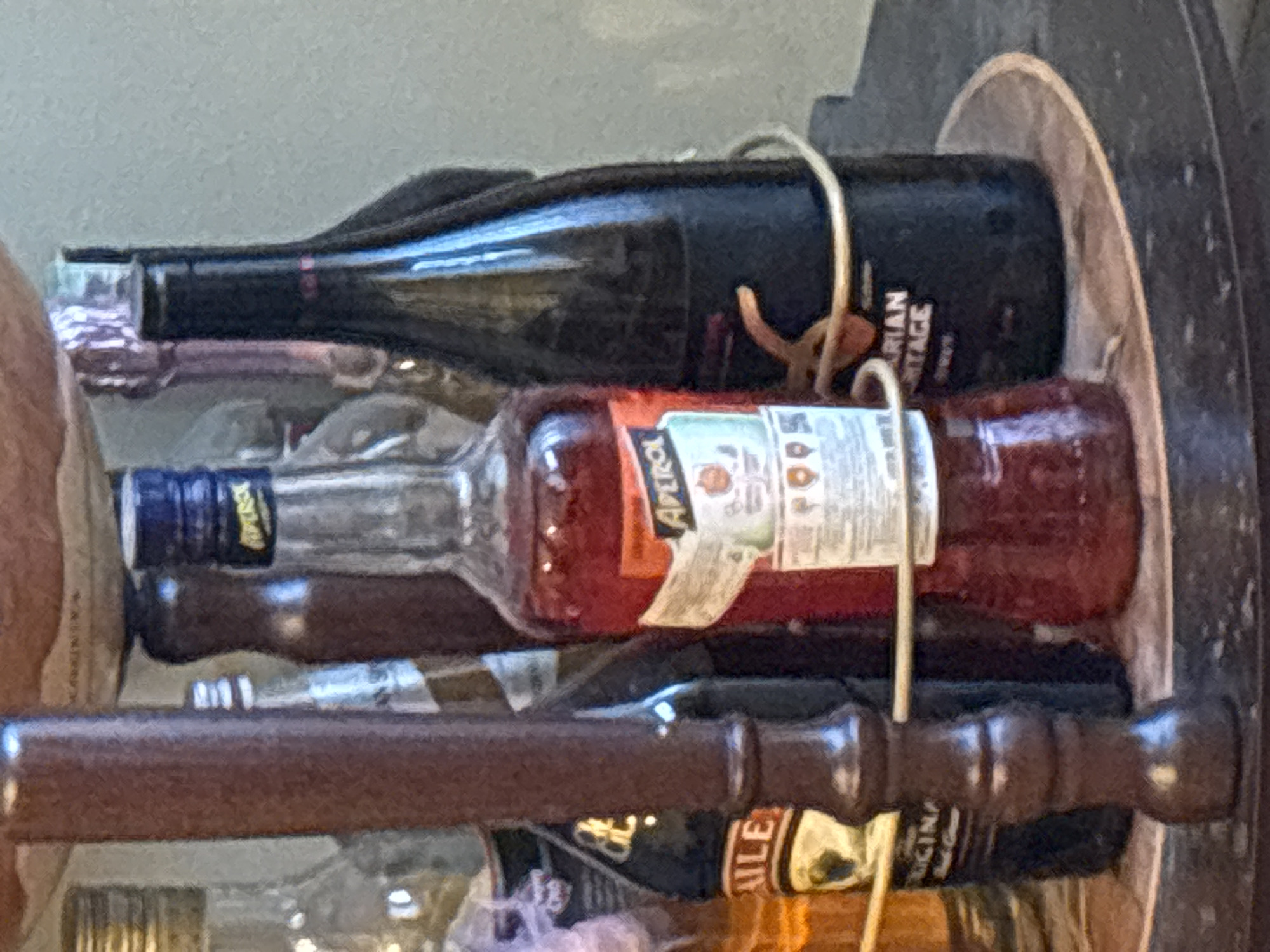 The image presents a close-up, horizontal view of several bottles of alcohol arranged in a storage rack, likely part of a liquor cabinet or home bar. The bottles are held in place by curved metal wires.
At the top, a dark-colored bottle, possibly wine, displays a label with the partial text "ARIAN HERITAGE" and a decorative element. Directly below it is a clear glass bottle containing an orange liquid, unmistakably identified as "APEROL" from its white label featuring red and orange text and graphics. Further down, a third clear glass bottle with a dark blue cap is partially visible, with "ABSOLU" faintly readable on its label. At the very bottom, a dark bottle can be seen with "BAILEYS" discernible on its label.
In the foreground, a prominent dark brown, carved wooden rod or handle extends horizontally across the frame, partially obscuring the lower bottles. The background is a blurry, light gray, suggesting an indoor setting with ambient light. There is no specific activity taking place, and the scene is a static arrangement of bottles. While the location context is given as Plovdiv, Bulgaria, the image itself offers no specific visual cues to confirm this. The lighting suggests it is daytime.