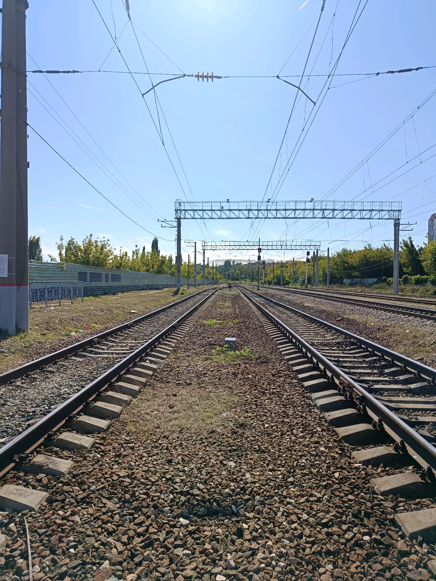 The image depicts a perspective view of a double set of railway tracks leading into the distance. The tracks are made of steel rails laid on concrete sleepers, covered in ballast composed of crushed stone. Overhead power lines are visible, suspended from poles and a large gantry structure. The sky is clear and blue, indicating daylight. The surrounding environment includes vegetation, a fence, and some distant buildings. There are no people or animals visible in the frame. The scene appears to be an operational railway yard or a section of track on the outskirts of a city. The specific location in Saratov, Russia, is not visually identifiable from the image alone.