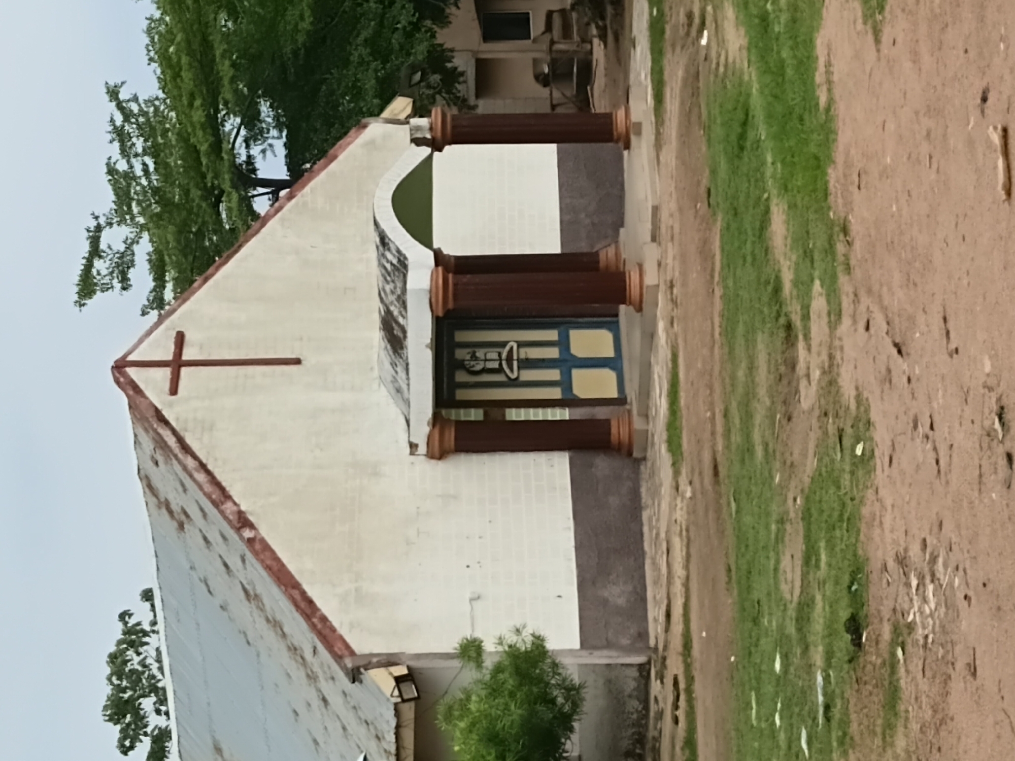 The image depicts the exterior of a modest church building, likely located in the city of Jalingo, Nigeria, based on its architectural style and surrounding environment. The building features a white facade with a dark grey band along the bottom. A prominent red cross adorns the triangular gable. The roof appears to be corrugated metal, showing signs of wear and tear. A small portico extends from the front of the building, supported by four dark red columns with decorative brown tops and bases. The portico is topped with a green arched section. A blue and yellow door with a glass panel is visible beneath the portico. The ground in front of the church is uneven, with patches of green grass and dirt. Trees are visible in the background, and the sky is overcast. There are no people or animals visible in the frame.