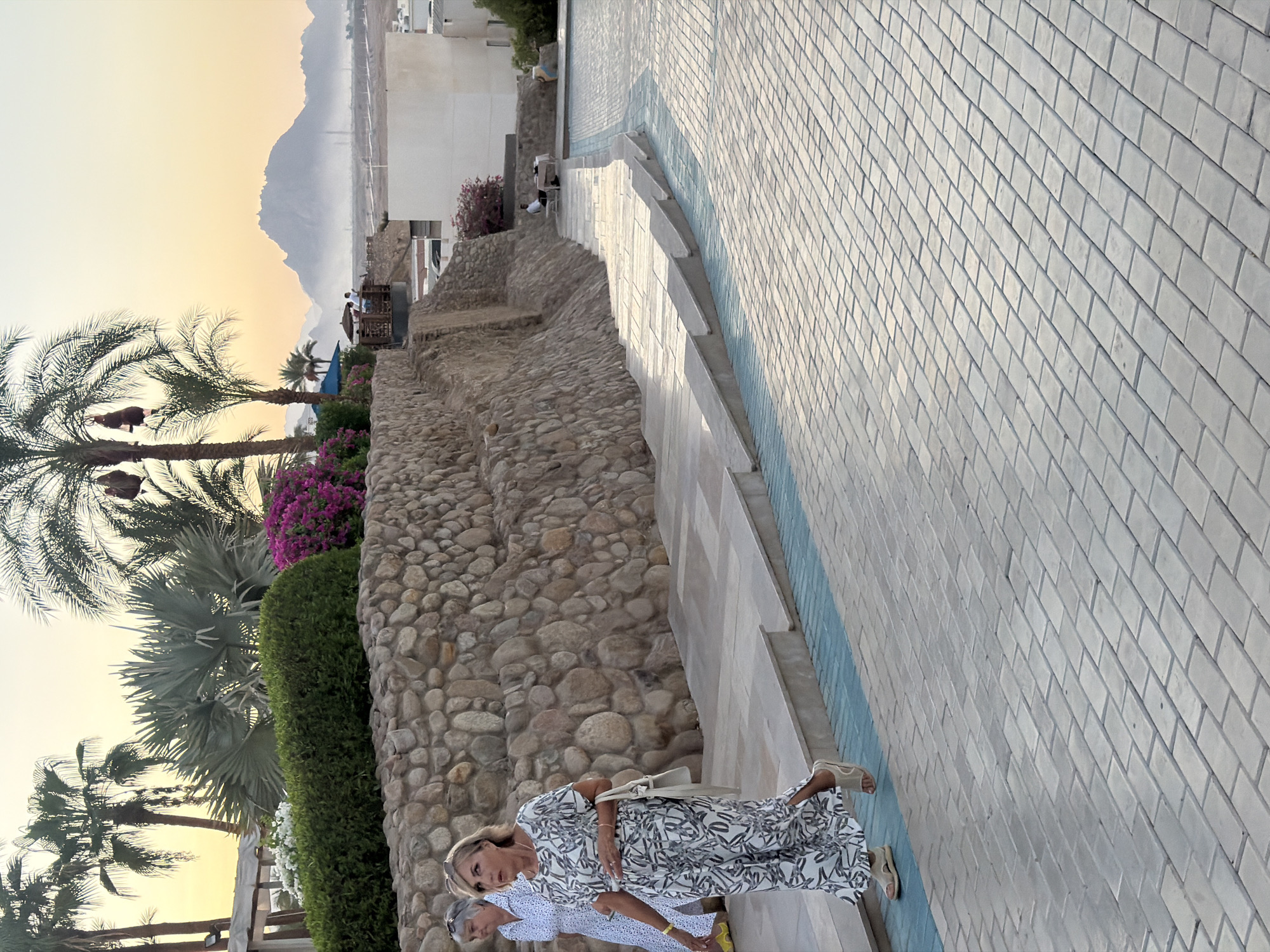 The image captures a scene in Sharm El Sheikh, Egypt, with a focus on an outdoor paved area and a stone wall. Two women are walking on a pathway. The woman in the foreground is wearing a long, patterned dress and sandals, and she is carrying a white purse. The other woman, slightly behind her, is wearing a white polka-dot dress. Behind the women, a textured stone wall forms a retaining wall, topped with lush green bushes and vibrant pink flowers. Palm trees and other tropical foliage are visible in the background. The sky is a soft gradient of yellow and orange, indicating either sunrise or sunset, with distant mountains shrouded in a light haze. A man is seated at a table in the far background, near a white building. The paved pathway is made of light-colored rectangular blocks, with a blue-striped border. The overall impression is a tranquil resort setting.