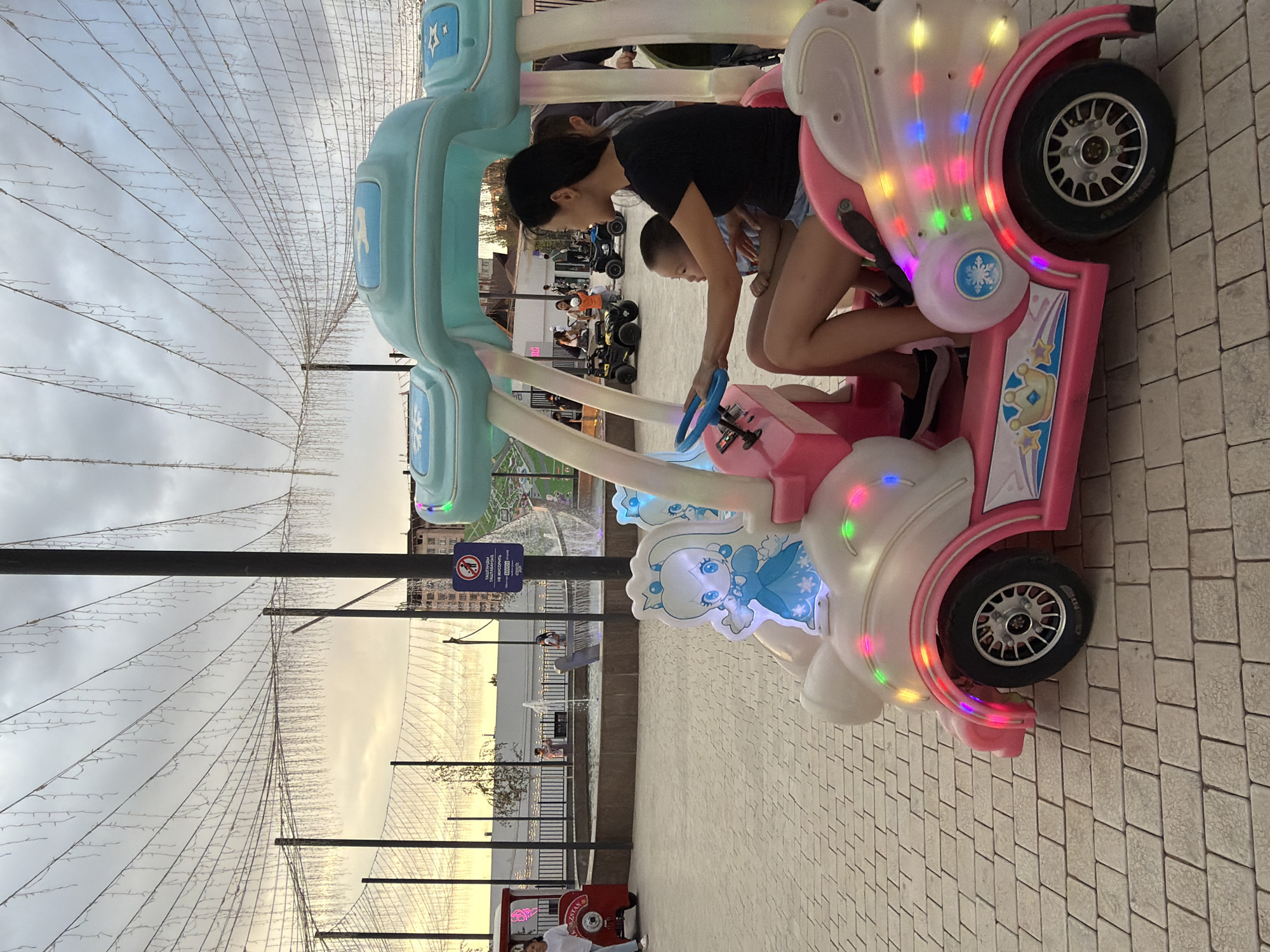 In an outdoor plaza paved with light brown bricks, a woman and a young child sit in a pink, cartoonish electric toy car. The car is decorated with blue and white accents, a cartoon princess graphic, and colorful LED lights. The woman, wearing a black t-shirt and denim shorts, is in the driver's seat, holding a blue steering wheel. The child, in a light blue shirt, sits in her lap.
In the background, several other children are riding similar toy vehicles. Beyond them, a fountain sprays water, and in the distance, buildings and trees are visible under a cloudy sky with warm tones. Strings of white lights are strung across the plaza, adding to the festive atmosphere. A lamppost with a warning sign is positioned to the right of the main subjects. A small red toy train is visible to the left of the frame.